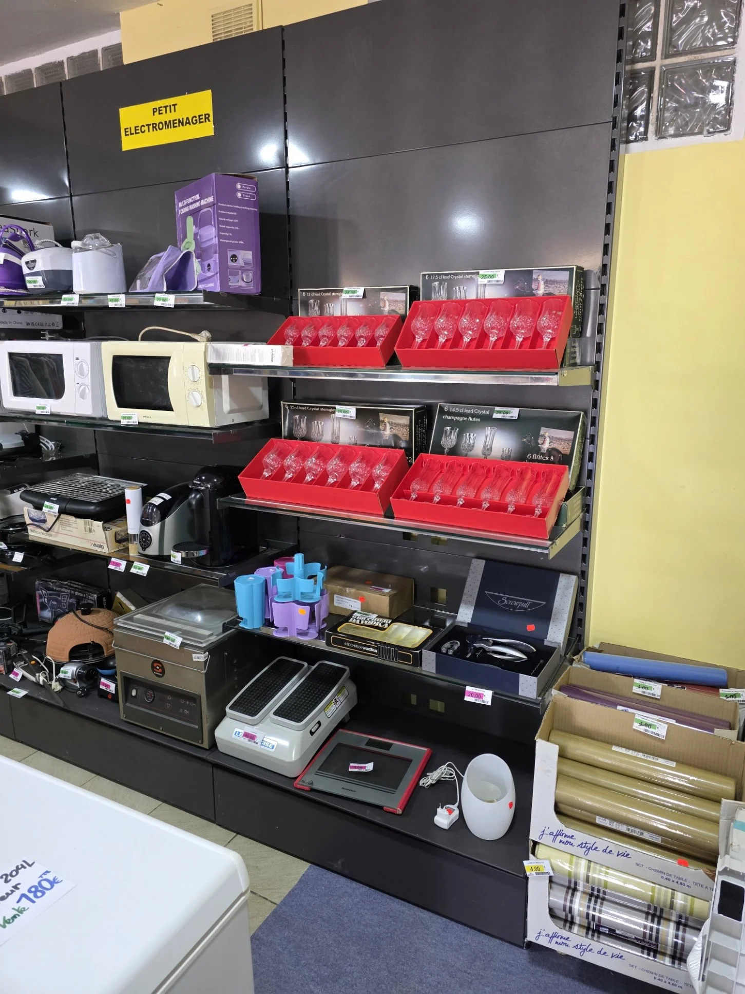 Une étagère dans un magasin contenant divers appareils électroménagers et objets, notamment des micro-ondes, des appareils à raclette, des ciseaux, un grille-pain, des tapis de sol, et des pièces de décoration comme des verres en cristal. Des étiquettes de prix sont visibles sur certains produits.