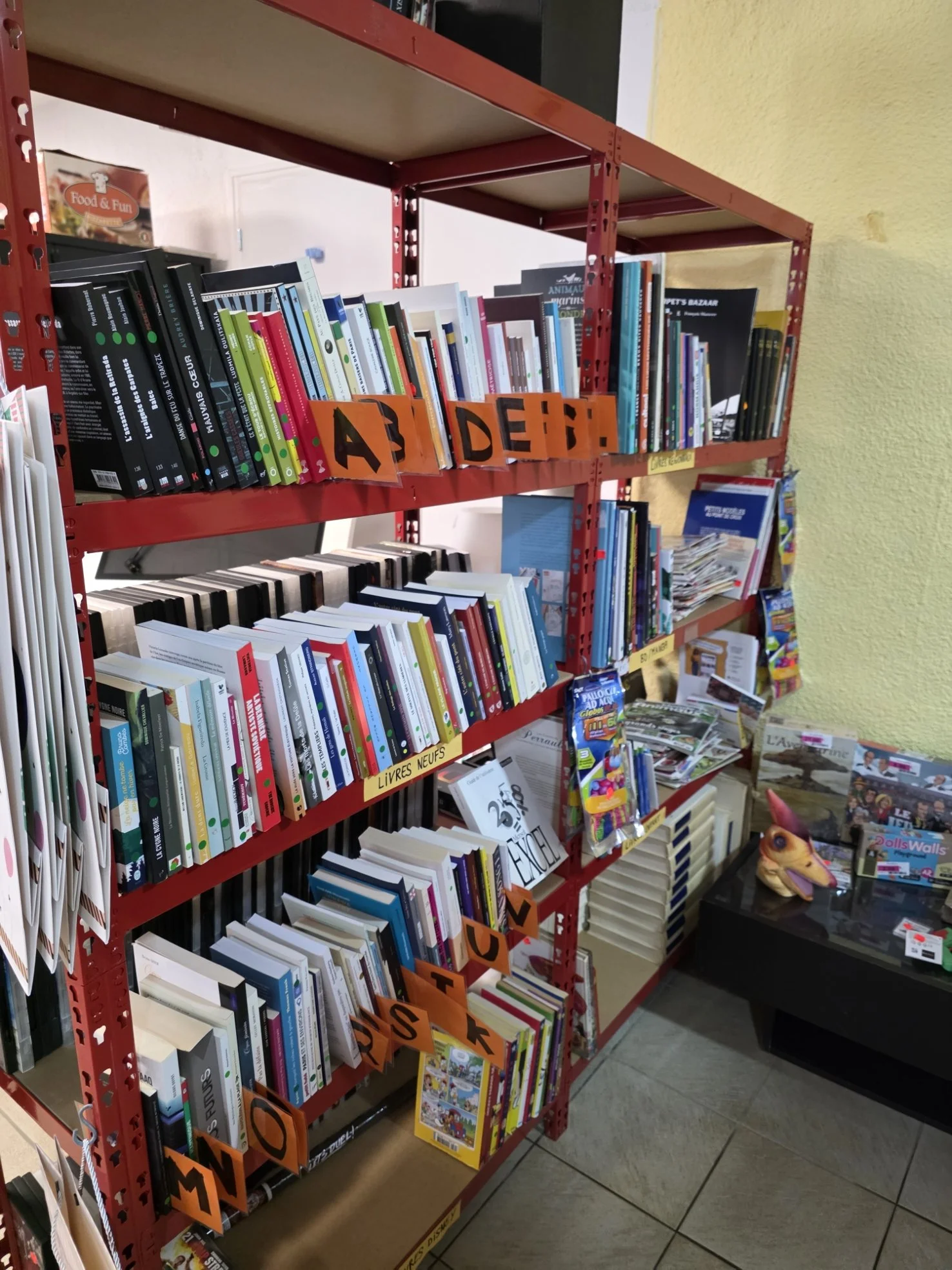 Étagère rouge contenant des livres dans une librairie, avec des signalisations en orange indiquant 'A', 'D', 'O', 'N', et 'Livres Neufs'.