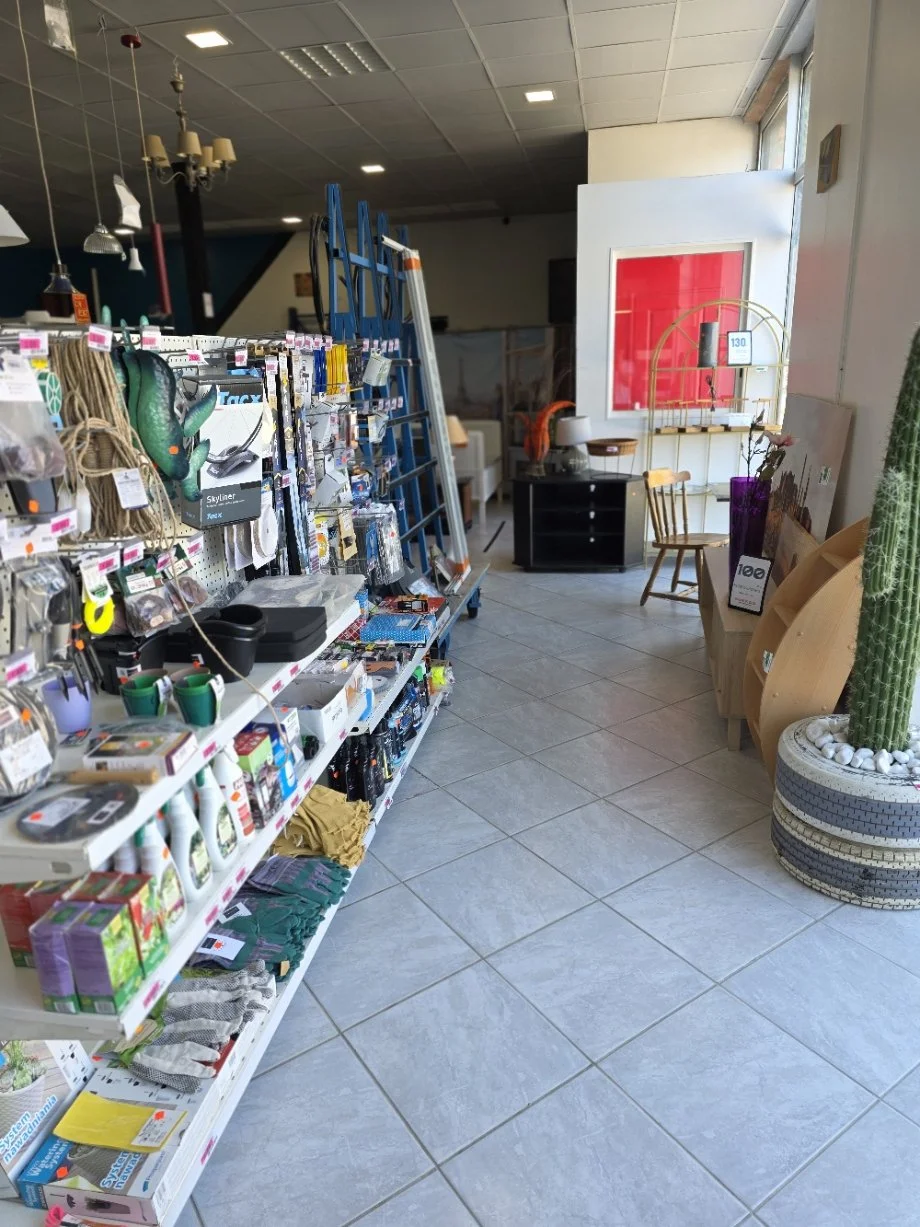 Intérieur d'un magasin avec des étagères remplie d'outils, d'accessoires, un escalier bleu, et des meubles, avec un coin décoration en arrière-plan.