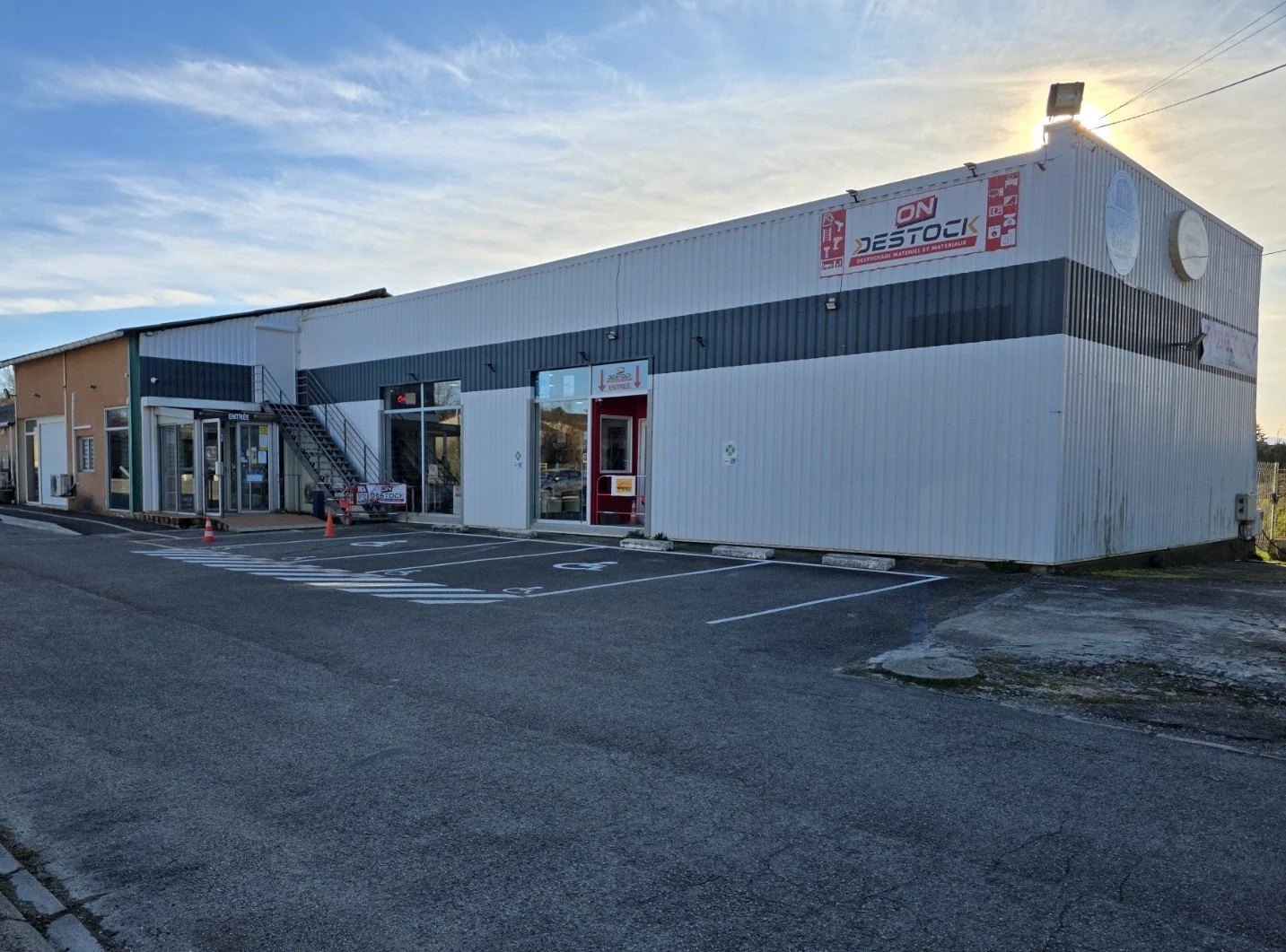 Un bâtiment commercial avec un parking vide devant, des panneaux publicitaires, un escalier extérieur et des fenêtres, sous un ciel en fin de journée.