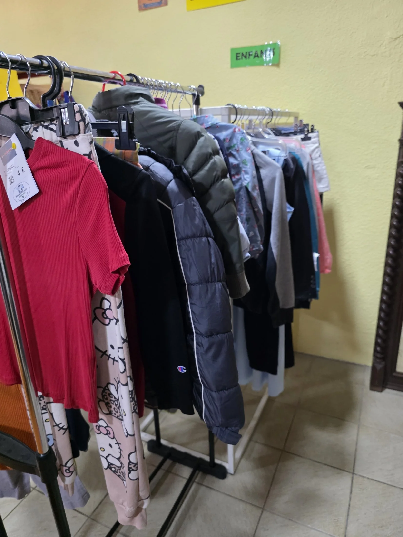Vêtements pour enfants suspendus sur une étagère dans une boutique, avec un panneau vert indiquant 'Enfants' au mur en arrière-plan.