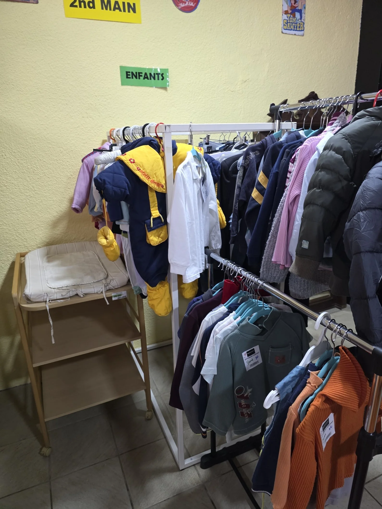 Vêtements pour enfants et adultes suspendus sur des portants dans un magasin d'occasion, avec signes indiquant '2nd Main' et 'Enfants'.