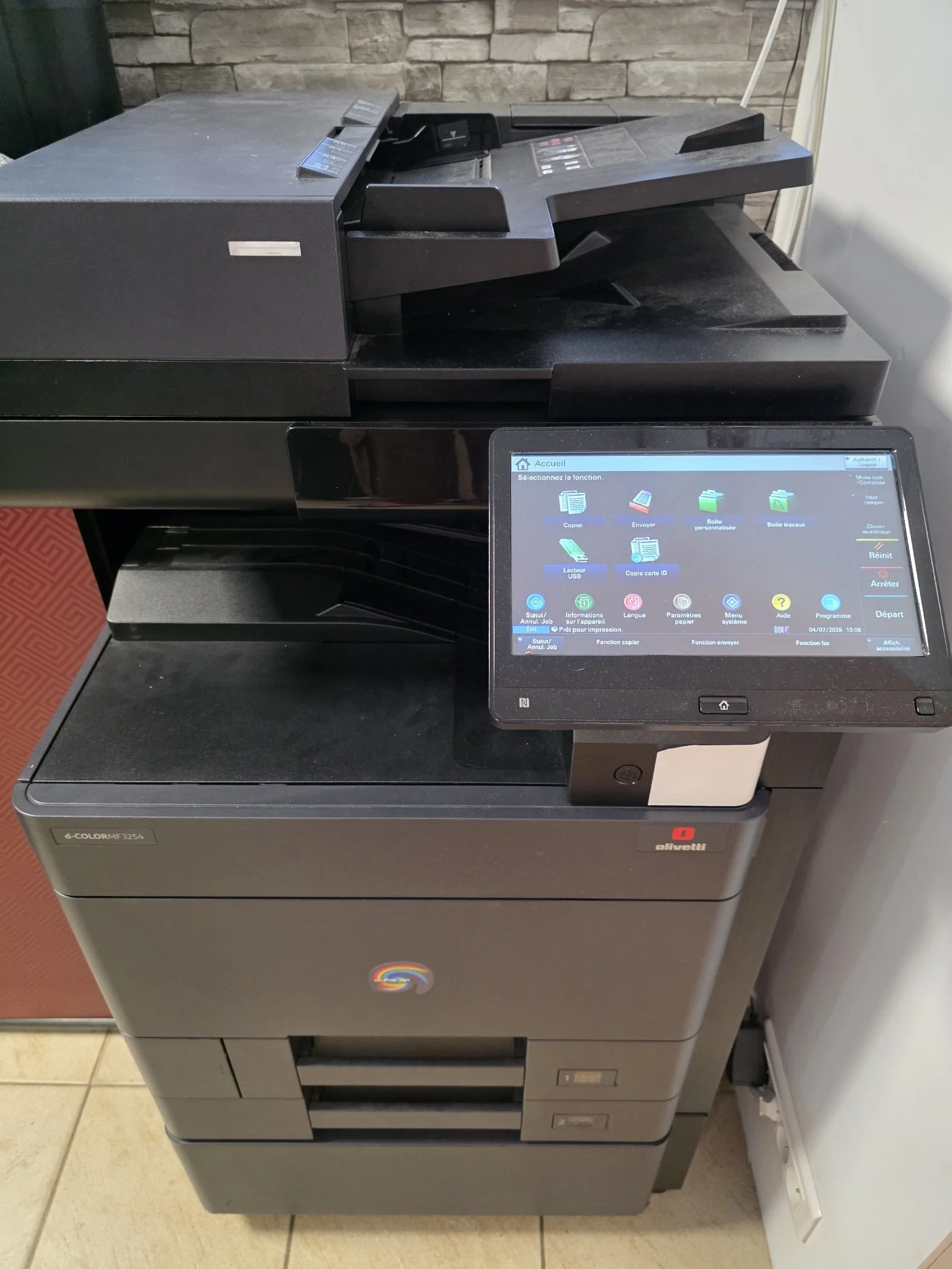Imprimante multifonction noire avec écran tactile, située à côté d'une prise électrique sur un mur. La marque 'Olivetti' est visible sur la machine.