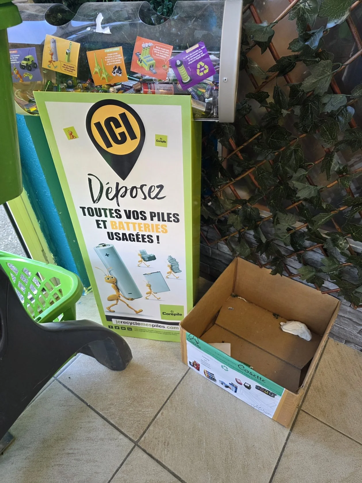 Un panneau publicitaire invitant à déposer les piles et batteries usagées dans une boîte de recyclage. La boîte est vide, avec une illustration d'une pile anthropomorphe. À côté, une boîte en carton vide et un panier vert à gauche.