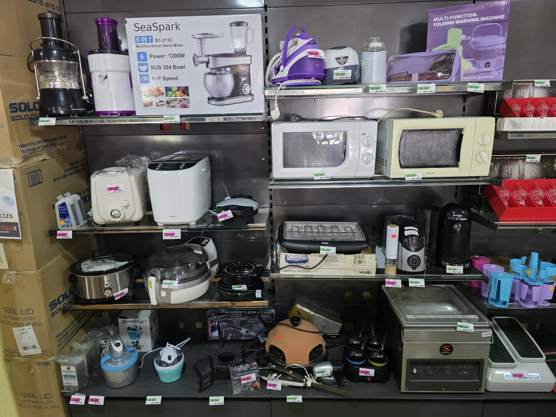 Étagère de divers appareils ménagers et accessoires, incluant des mixeurs, micro-ondes, grille-pain, et autres petits appareils électriques avec étiquettes de prix.