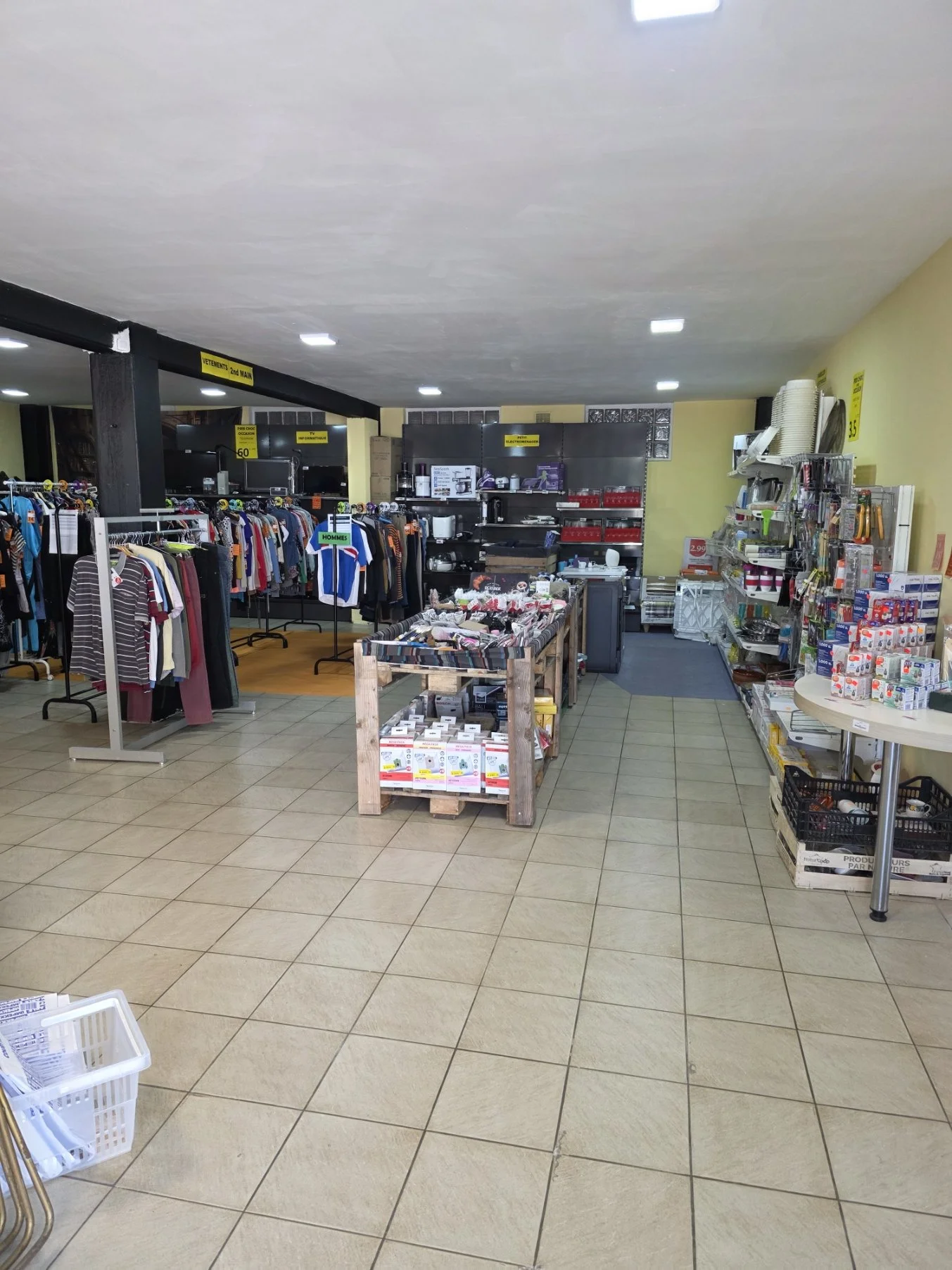 Intérieur d'un magasin avec des vêtements suspendus, des étagères avec divers articles et un sol en carrelage.