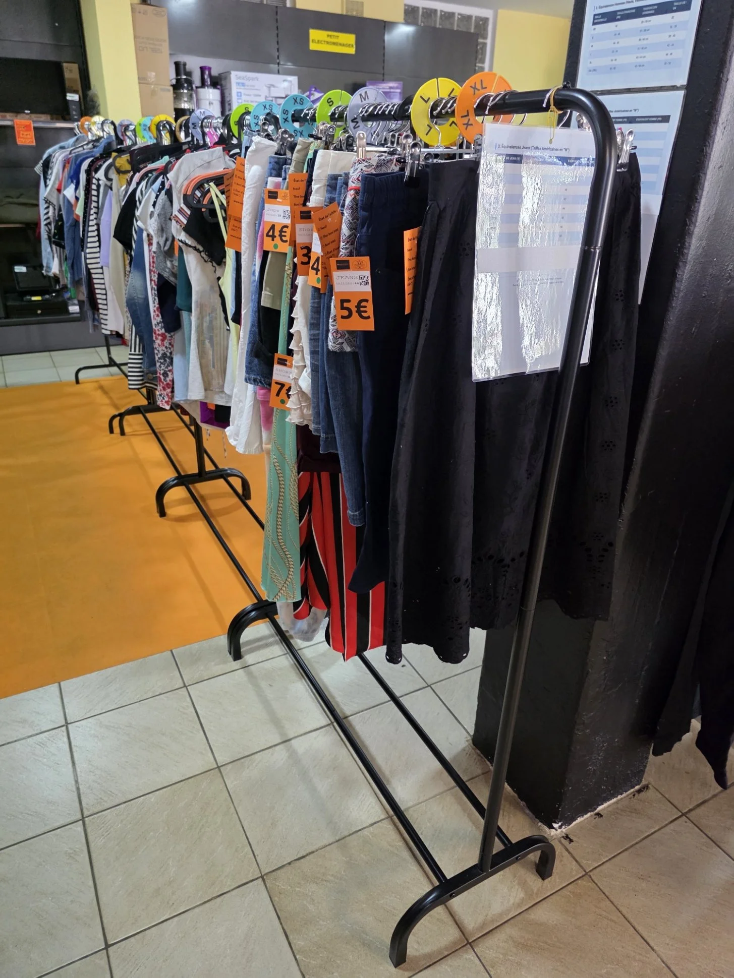 Rack de vêtements dans un magasin de seconde main, avec des vêtements pour femmes et hommes, tags de prix orange, et une affiche en arrière-plan indiquant des tailles.