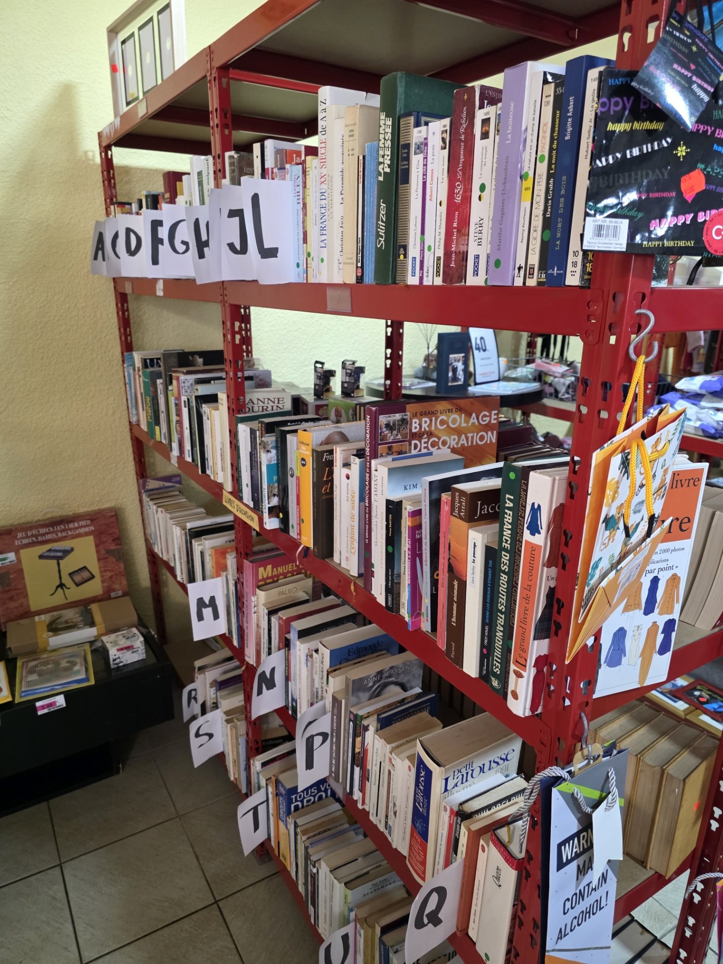 Rayonnage rouge rempli de livres en français, avec des étiquettes en forme de lettres et des cartons suspendus avec des illustrations, dans une librairie ou un vide-grenier.