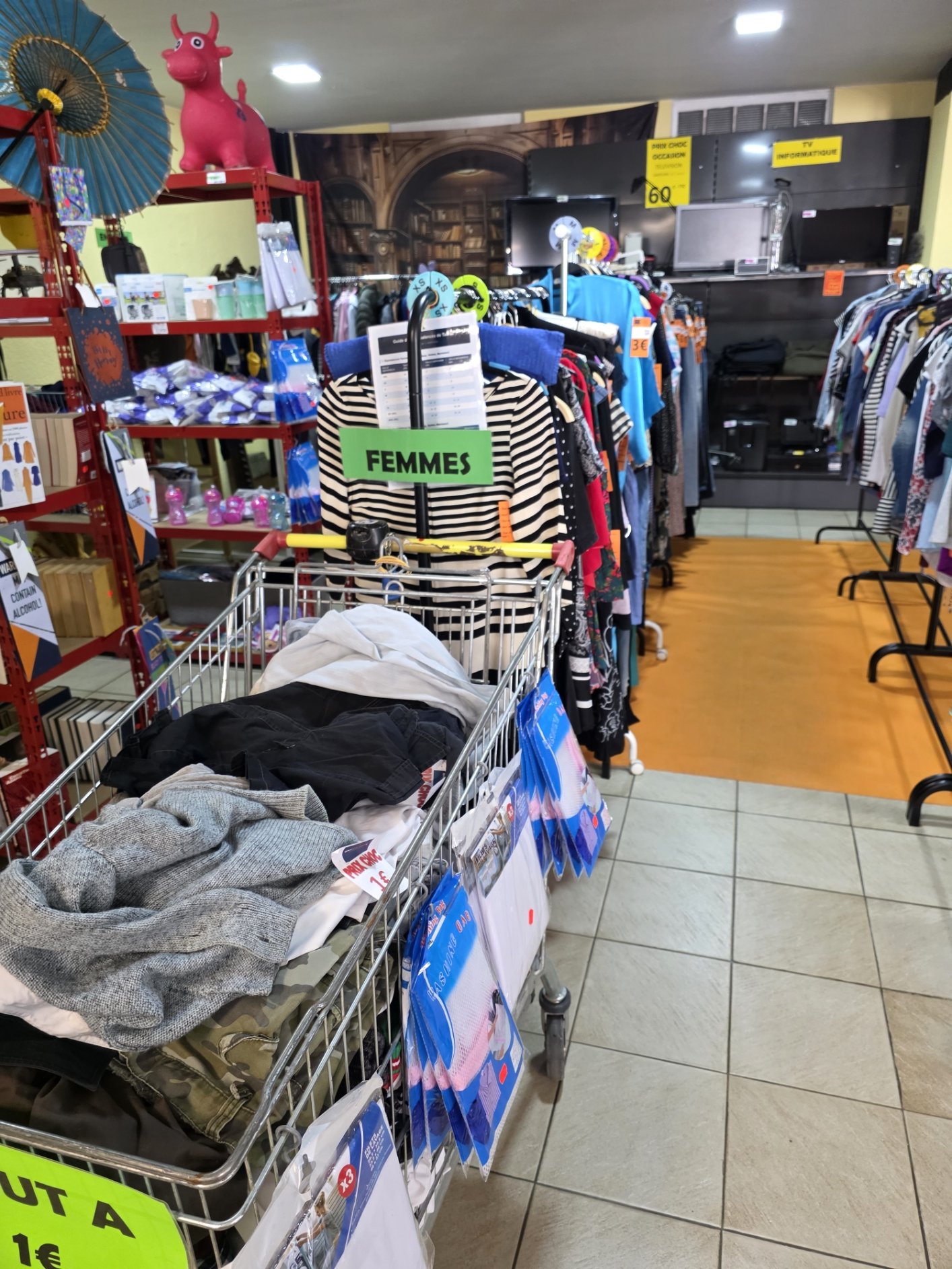 Une photo de rayon d'un magasin d'occasion avec des vêtements pour femmes en premier plan, une chariot contenant des vêtements, une étagère rouge avec des livres et des objets, un décor mural avec une bibliothèque en arrière-plan, et des panneaux jaunes indiquant la section informatique et la sélection. Deux jouets, un parasol et une figure de vache rose, sont sur une étagère en haut.