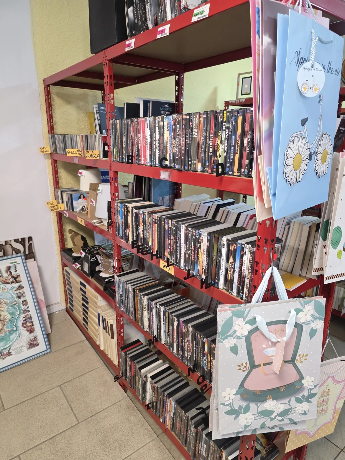 Rayonnage rouge avec CD, DVD et livres, plusieurs sacs cadeaux suspendus, section de magasin avec livres et décorations.