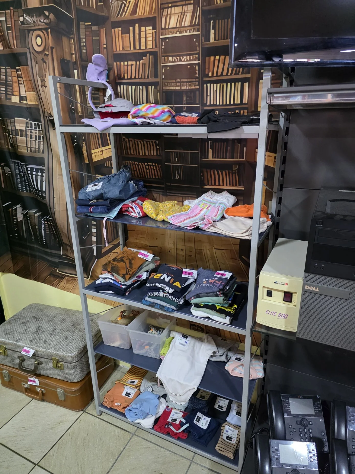 Une étagère métallique dans une boutique contenant des vêtements pliés, notamment des sous-vêtements, des vêtements de sport, des vêtements en coton, et un casque à côté. À droite, il y a une caisse enregistreuse et un moniteur.