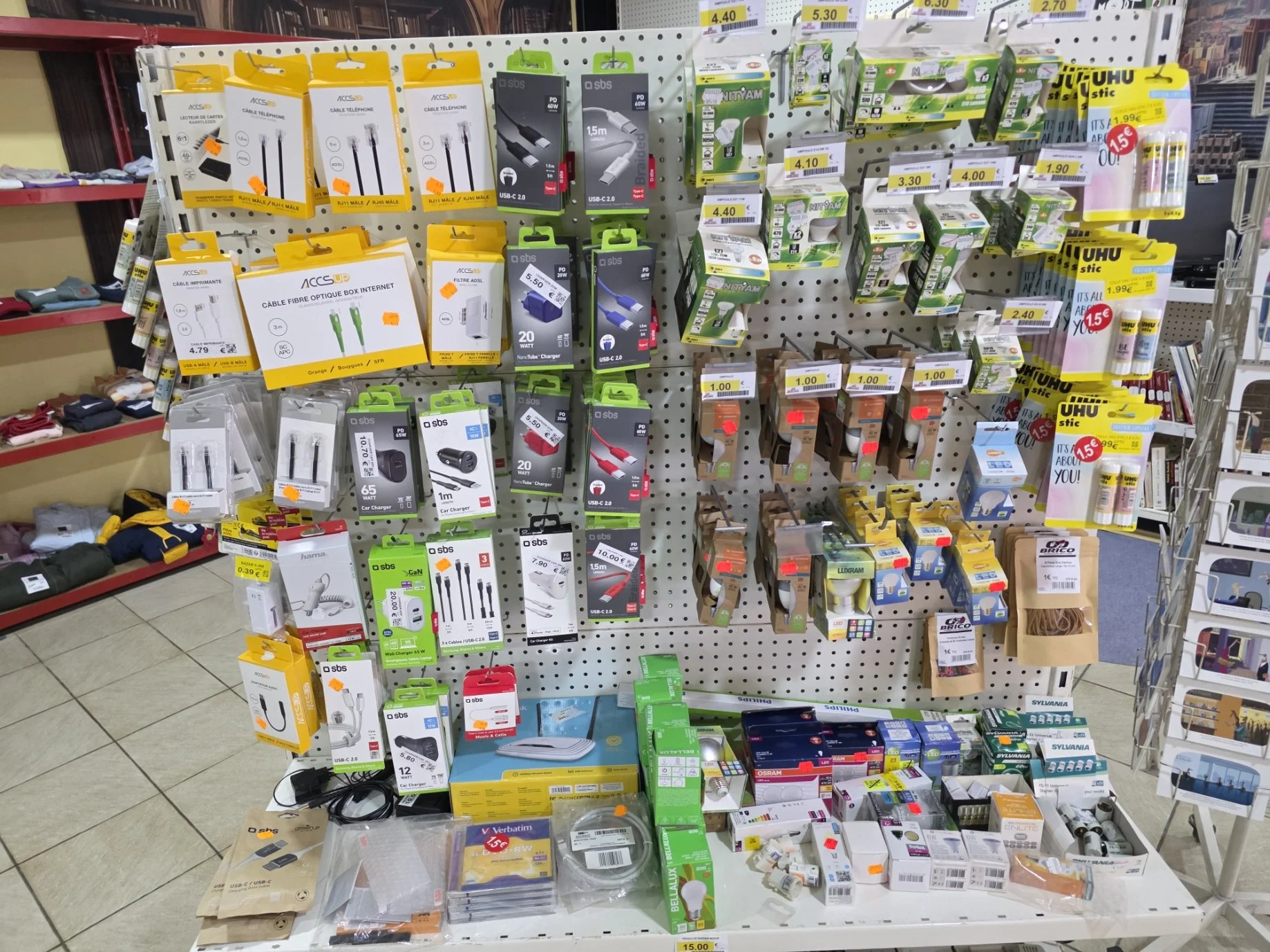 Vitrine avec divers câbles, chargeurs, ampoules, piles, ruban adhésif et petits accessoires électroniques. Disposition organisée en étagère de magasin.