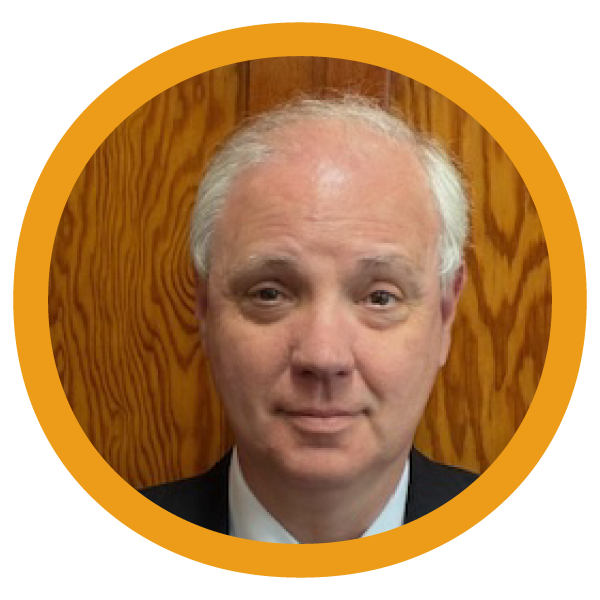 A portrait of a middle-aged man with light skin and white hair, wearing a dark suit and a white shirt, standing in front of a wooden wall.