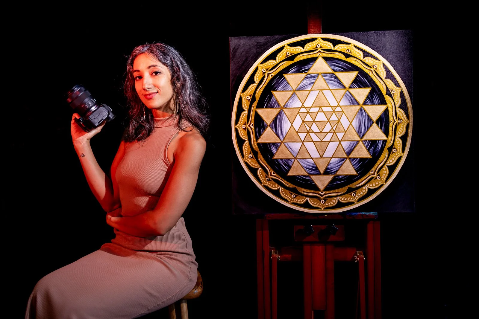 An artist holding a camera, sitting next to a large geometric artwork with gold and black patterns.