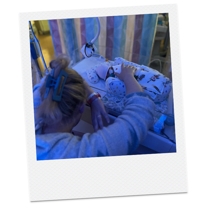 A person with blonde hair in a ponytail, wearing a gray sweater, is touching a sleeping baby in a hospital incubator. The baby is dressed in white and blue clothing with airplane prints, and is connected to medical tubes and equipment. The incubator has colorful curtains and medical devices around it.