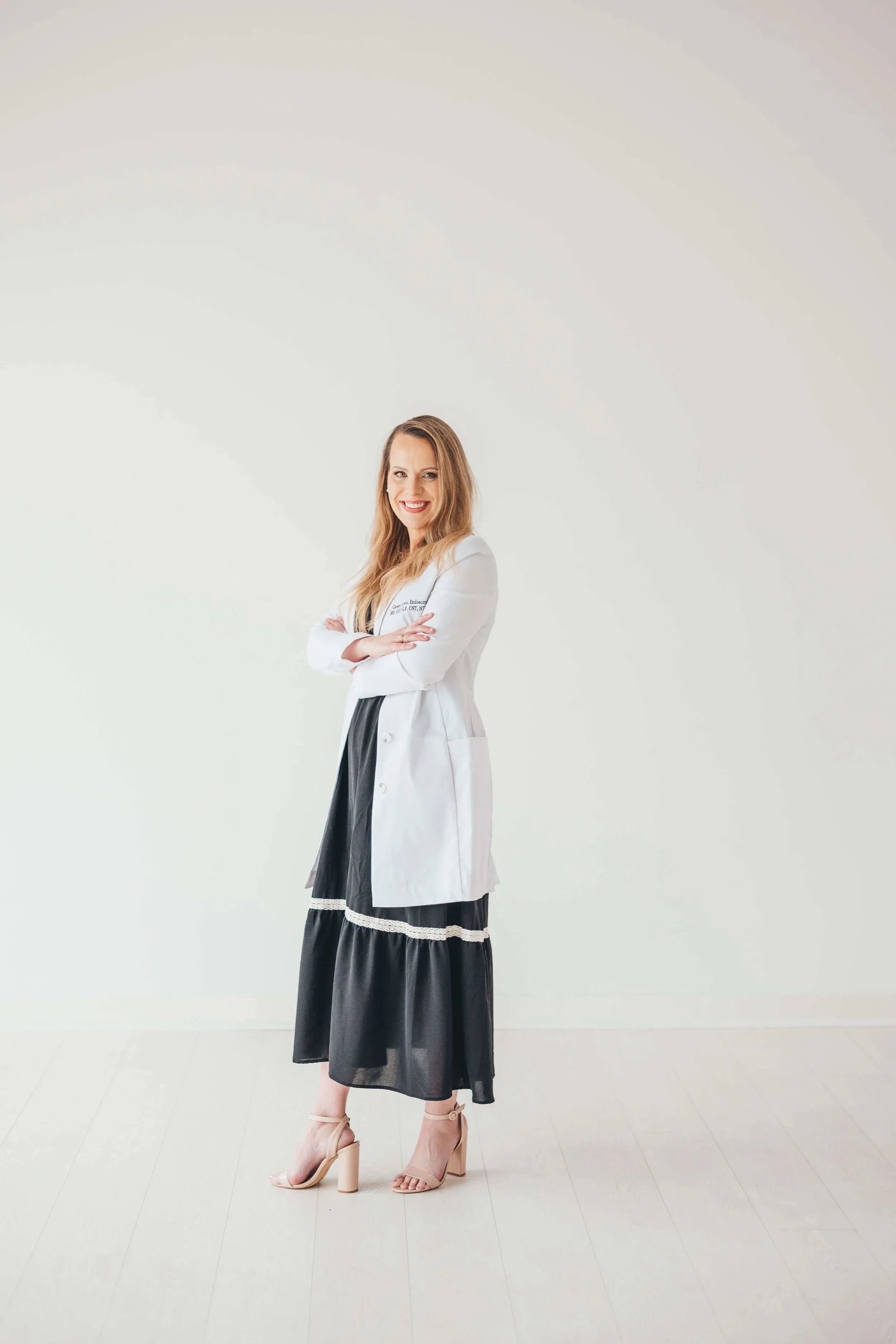 A woman smiling and posing with arms crossed, wearing a white blazer over a black dress with sheer overlay and high heels, standing against a plain off-white background.