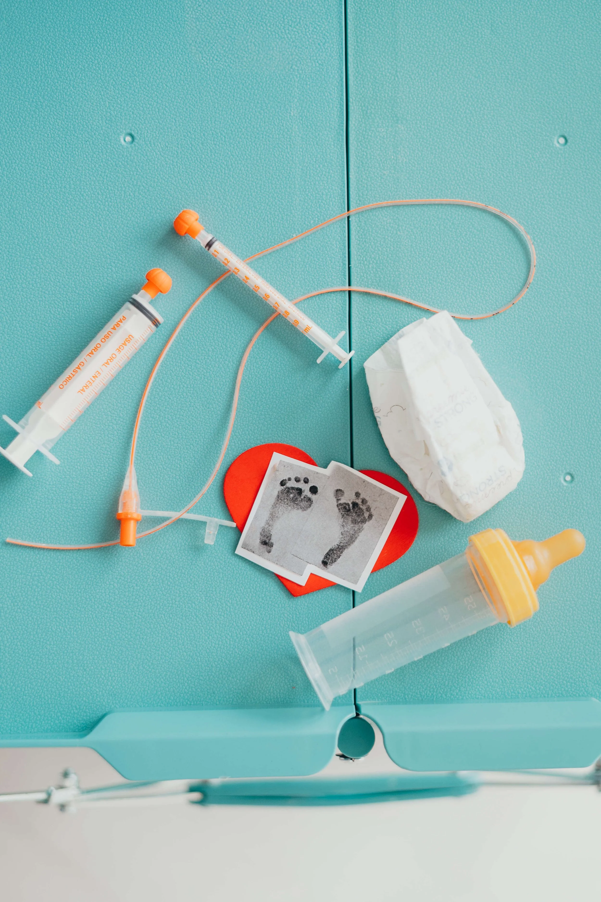 Medical supplies on a light blue tray including two syringes, a piece of tissue, a blood sample paper with footprints, and an empty specimen container.