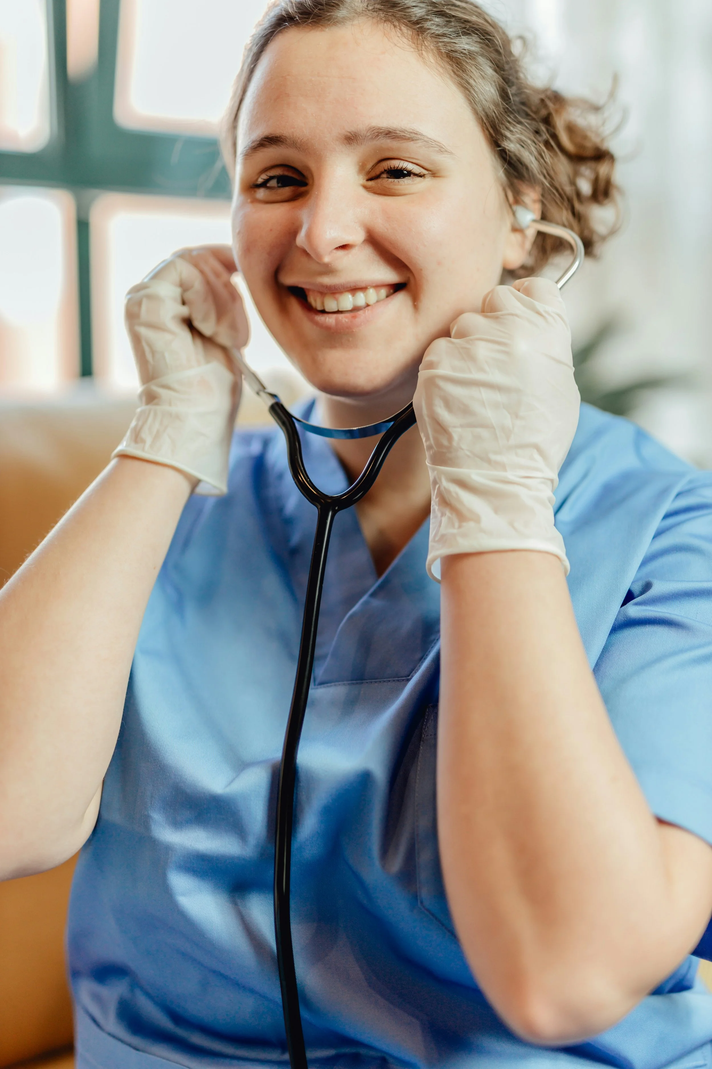 a young woman wearing blue medical scrubs and white gloves, smiling and adjusting a stethoscope around her neck in a bright, modern medical setting