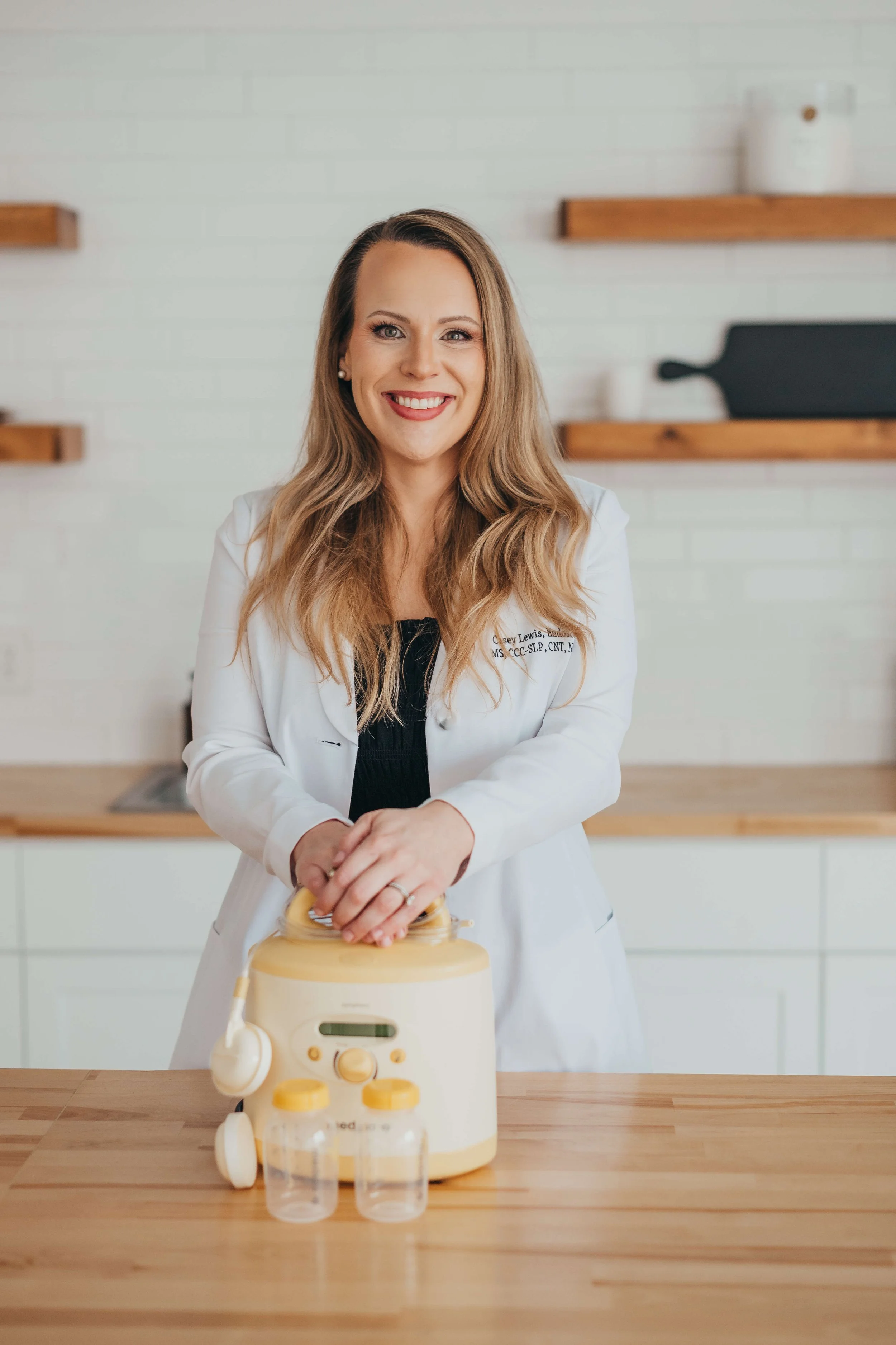 A woman wearing a white coat smiles at the camera, standing behind a wooden table with a Baby Essentials electric nebulizer and two clear plastic cups on it.