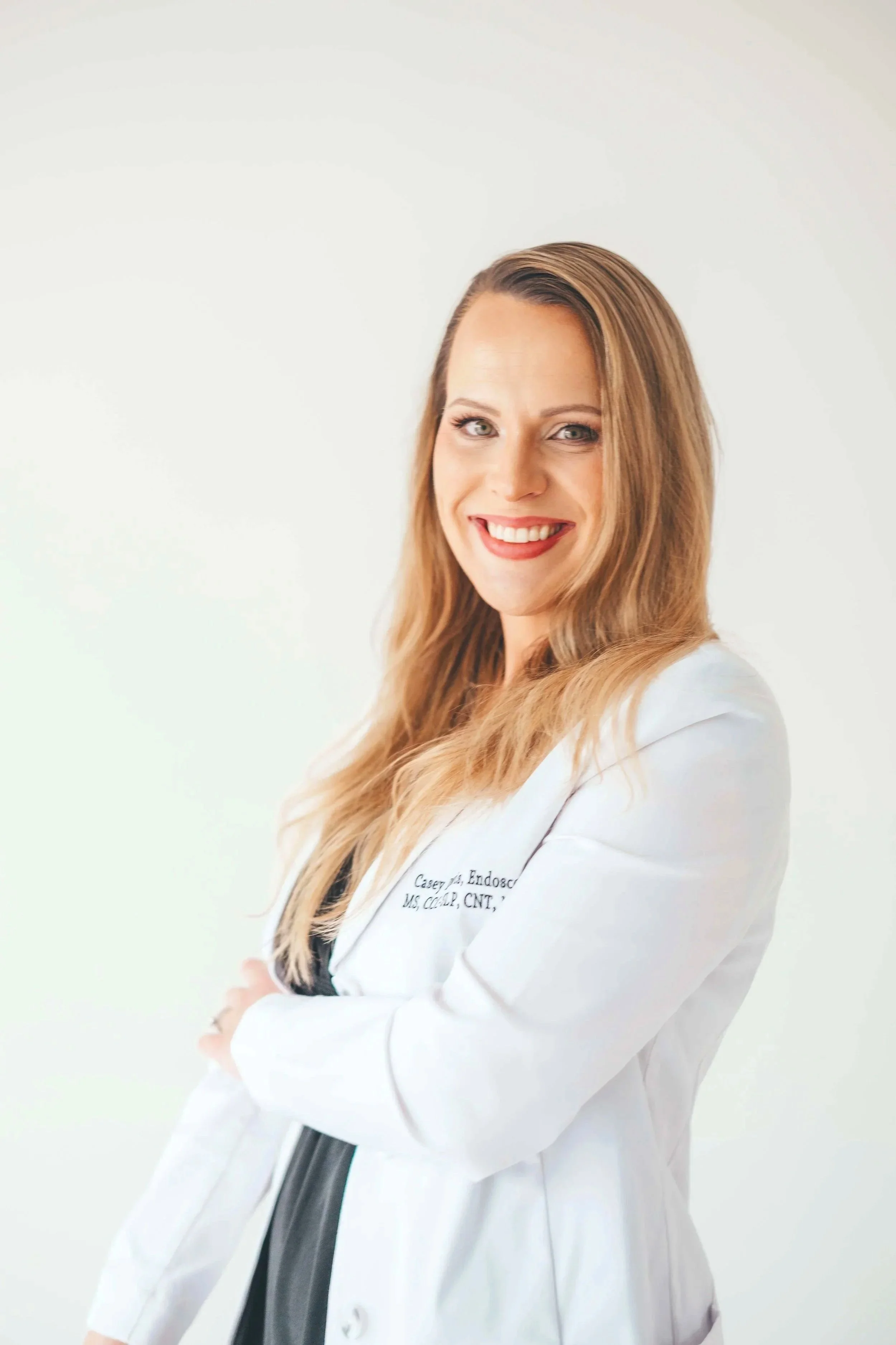 A woman with long blonde hair wearing a white medical coat smiling and standing against a plain light background.
