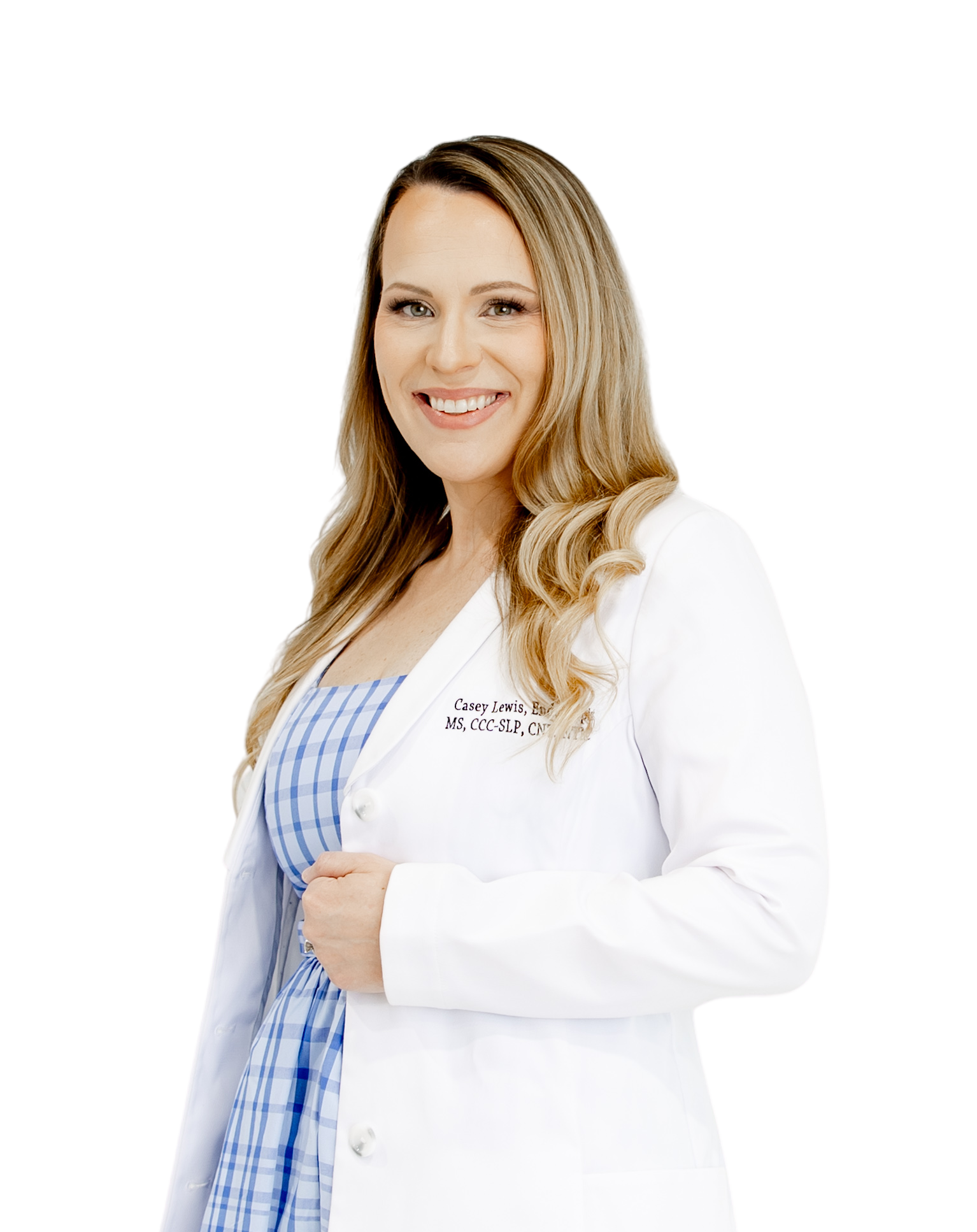 A smiling woman with long, wavy blonde hair wearing a white doctor coat over a blue and white checkered dress, standing with her hands clasped.