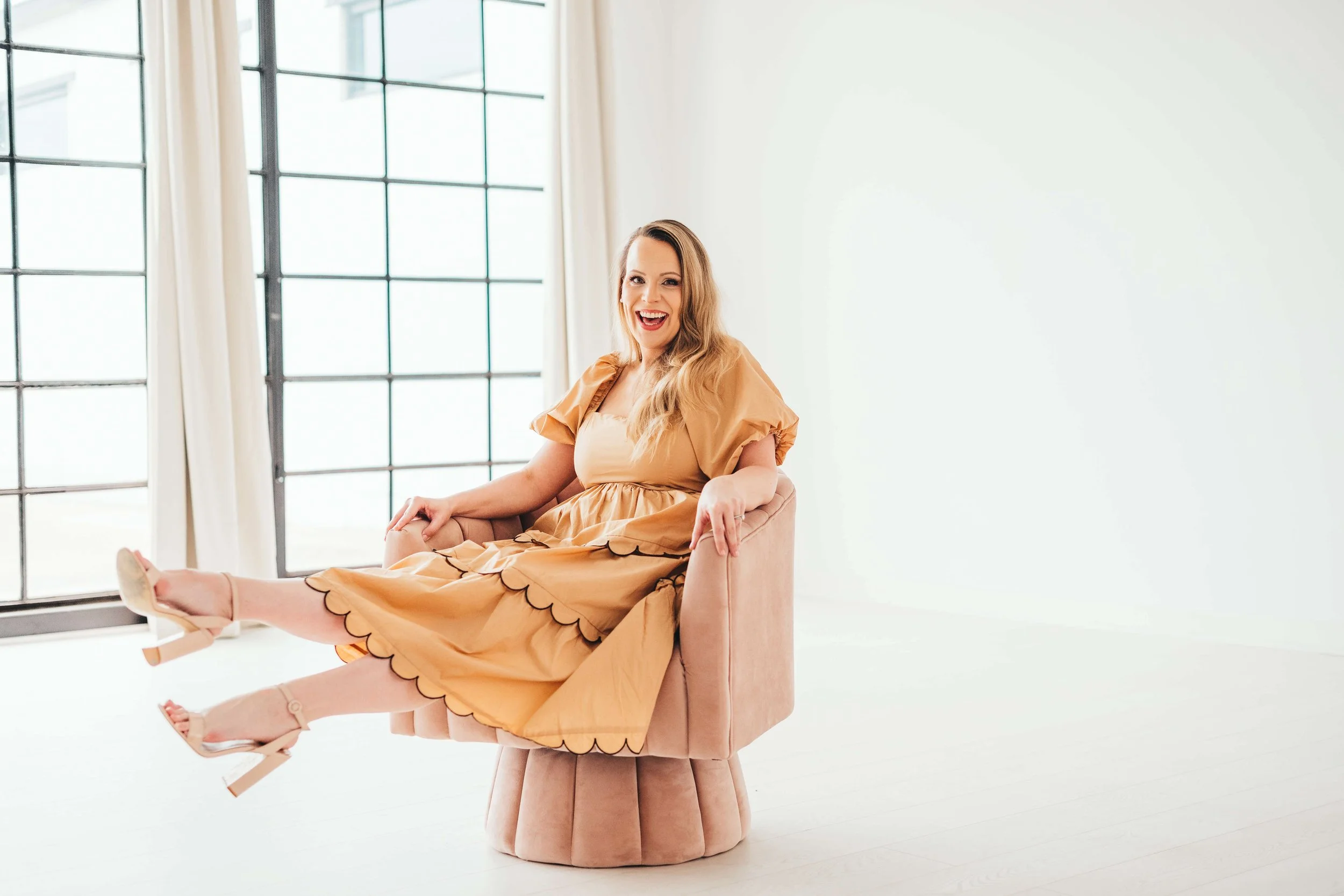 A woman with long, wavy blonde hair, smiling and sitting on a pink velvet chair with her legs extended. She is wearing a peach-colored dress with scalloped edges and beige high heels. The background features large window panes with white curtains, and the room has a minimalist and bright aesthetic.