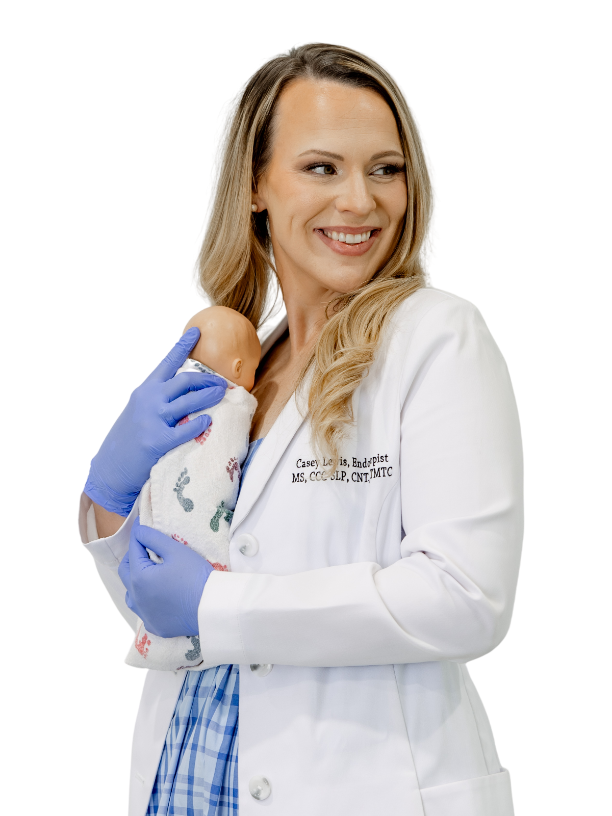 A woman in a white medical coat holding a newborn baby wrapped in a hospital blanket. The woman is smiling and looking to the side, wearing blue gloves, and has long blonde hair.