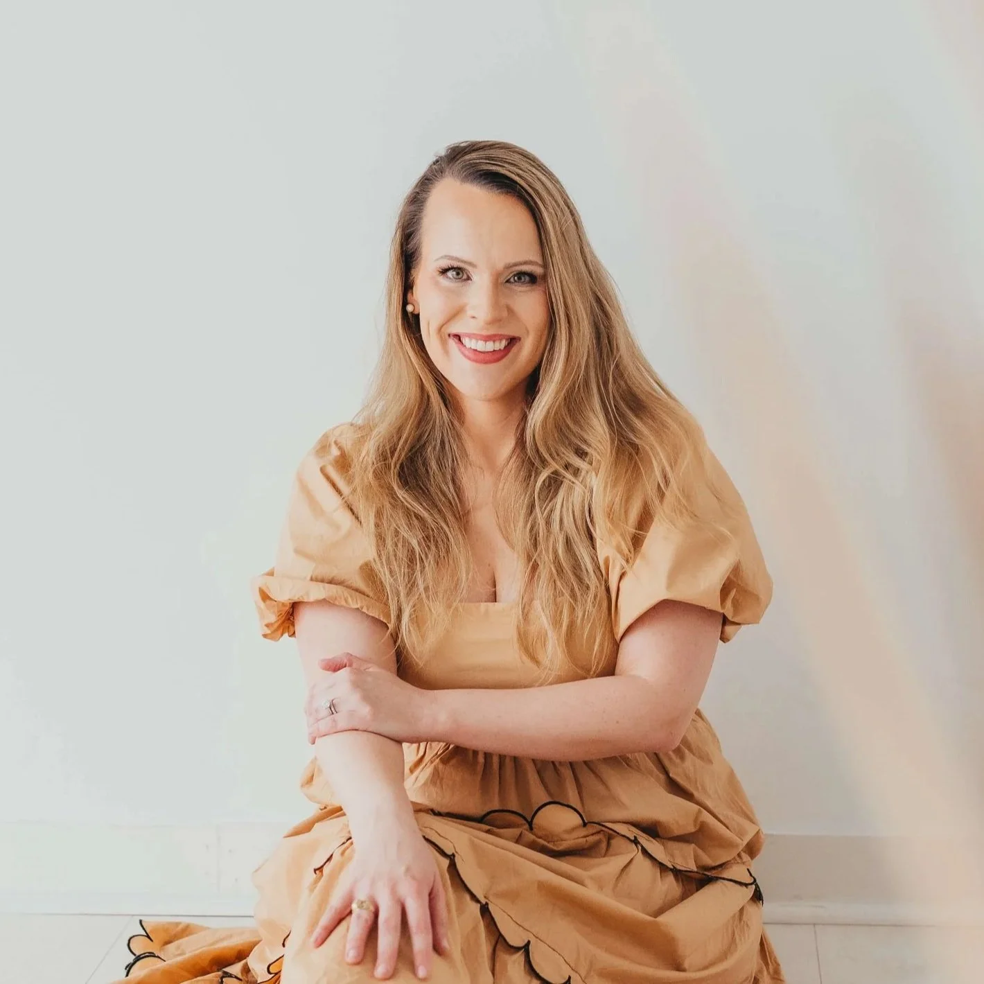 Woman with long wavy hair, wearing a beige dress with puffy sleeves, smiling, sitting on the floor against a plain light-colored wall.