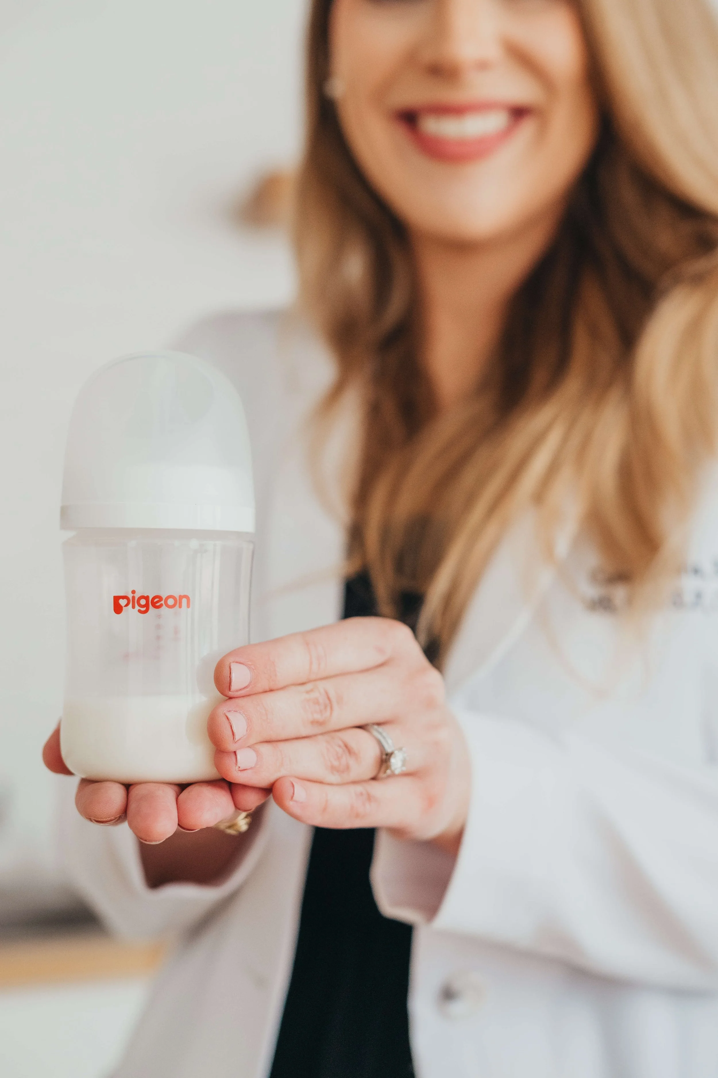 A woman with long wavy hair holding a Pigeon brand baby bottle with milk, smiling.