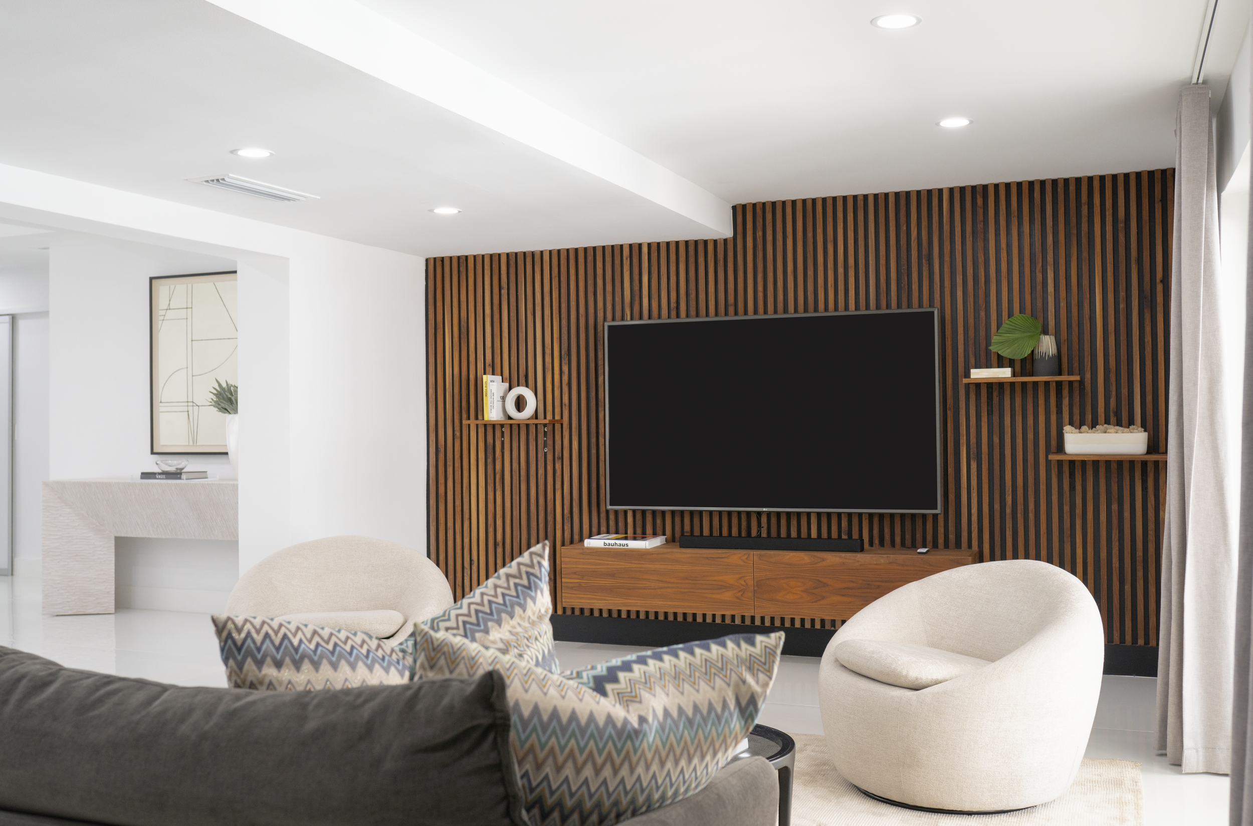 Modern living room with a wall-mounted flat-screen TV, wooden slat accent wall, and contemporary furniture including a patterned armchair, white rounded chair, and a dark sofa. Decor includes shelves with books and decorative items.