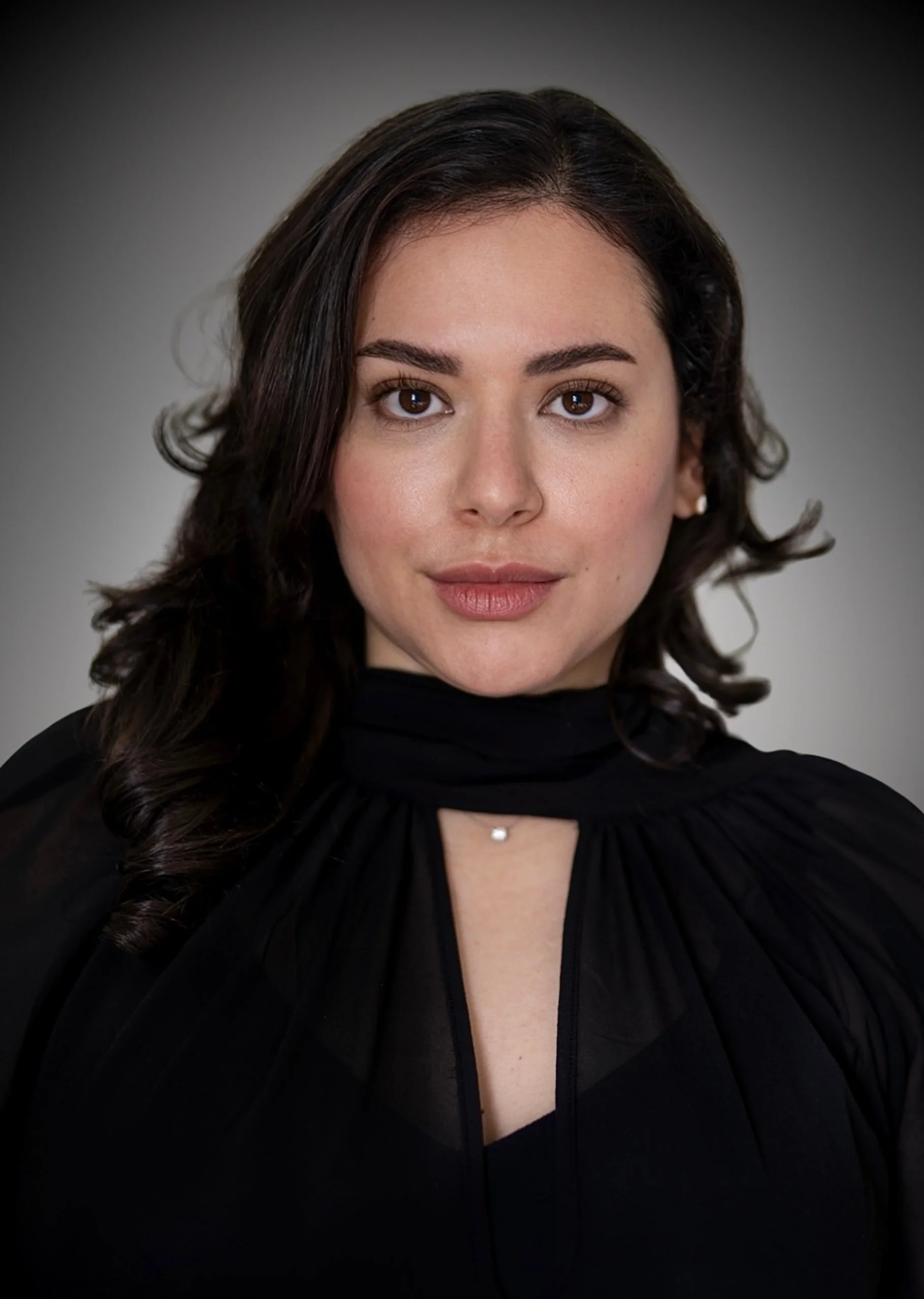 A woman with long, dark, wavy hair and brown eyes, wearing a black top with a high neck and a small necklace, looking directly at the camera against a plain gray background.