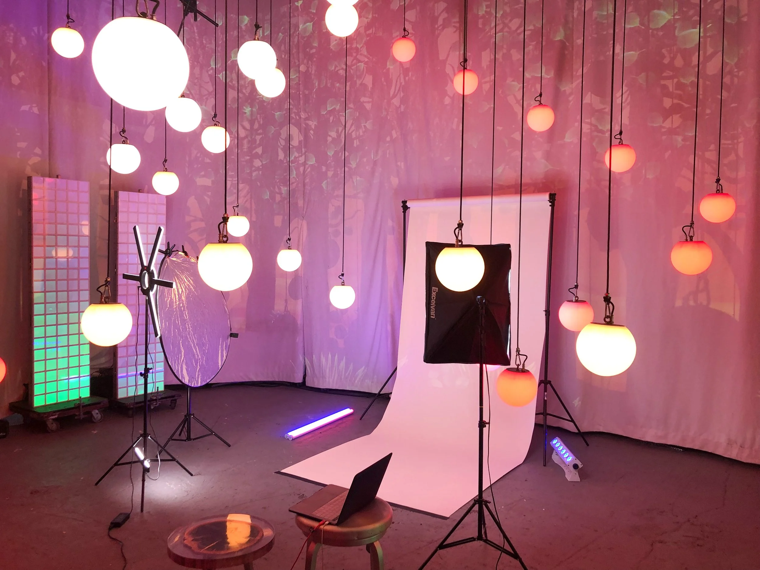 A photography studio setup with hanging spherical lights, a white backdrop, a softbox light, a laptop on a stool, and colorful lighting effects on the walls.