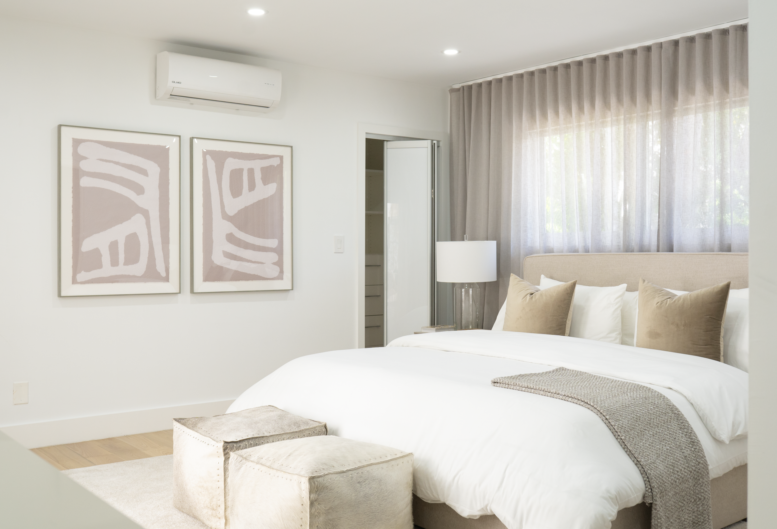 A bedroom with a large bed, beige headboard, white bedding, and beige pillows. A white lamp on a side table, window with sheer curtains, two abstract art pieces on the wall, and an air conditioning unit above.