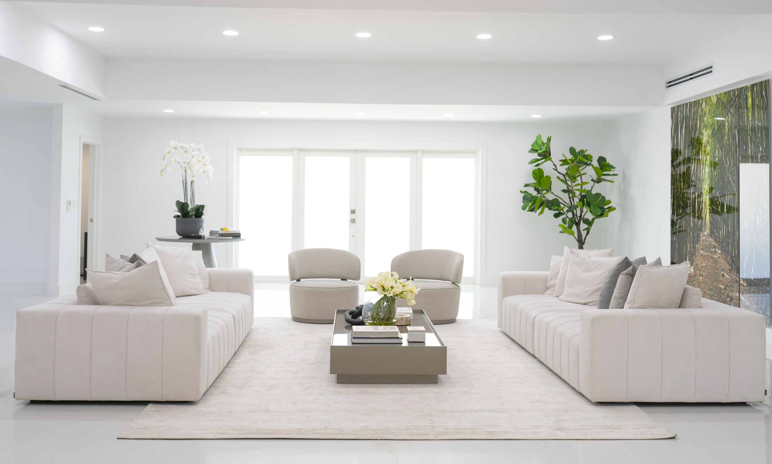 Bright, modern living room with two white sofas, a glass coffee table with flowers, two beige chairs, green plants, and a large wall mural of a forest scene.