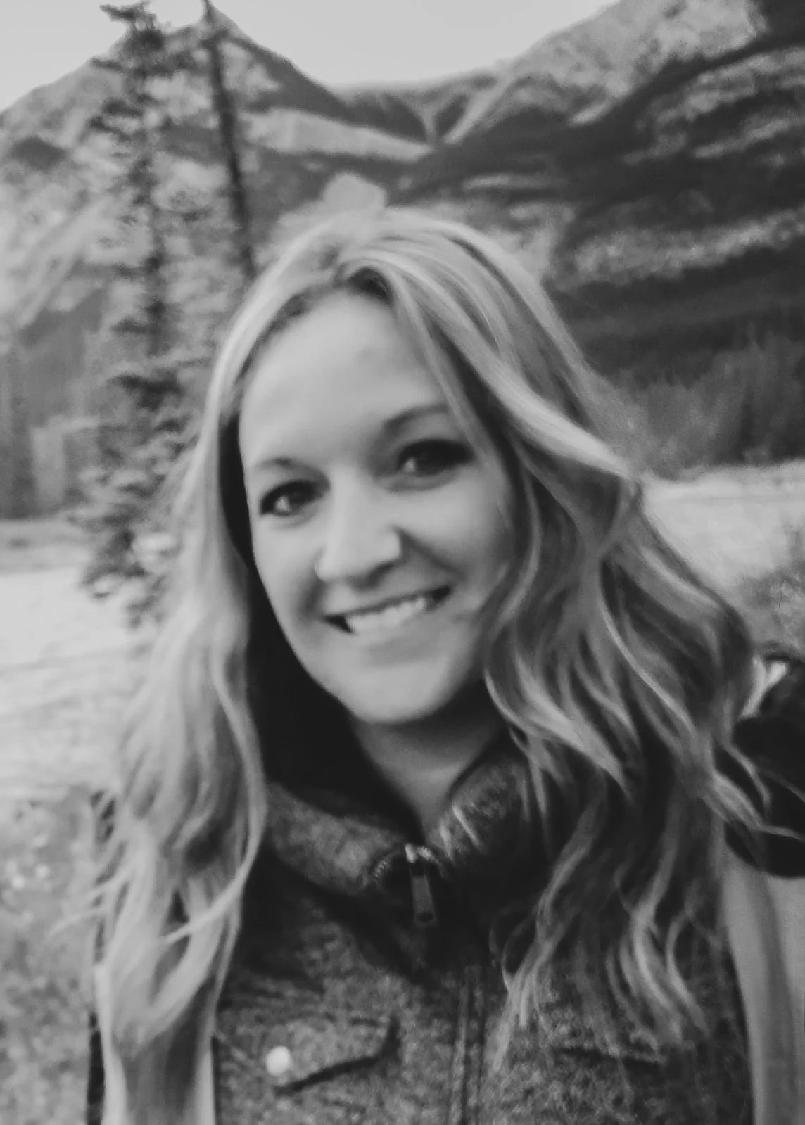 A woman smiling with long, wavy hair outdoors in front of a mountainous landscape with trees.