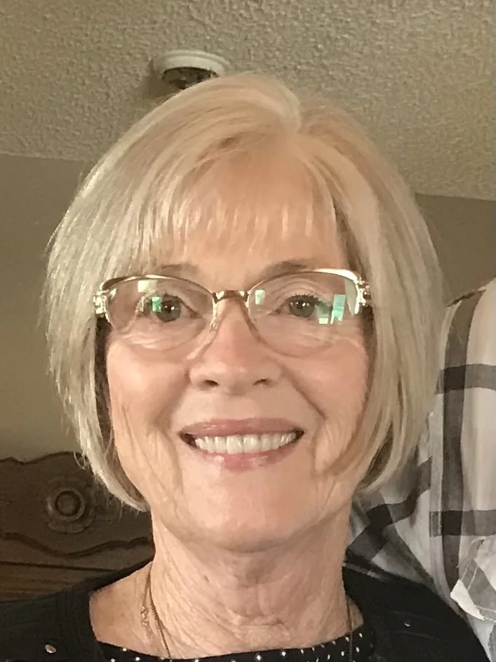 A smiling older woman with short blonde hair and glasses, wearing a black top with small white polka dots, a gold necklace, and a friendly expression.