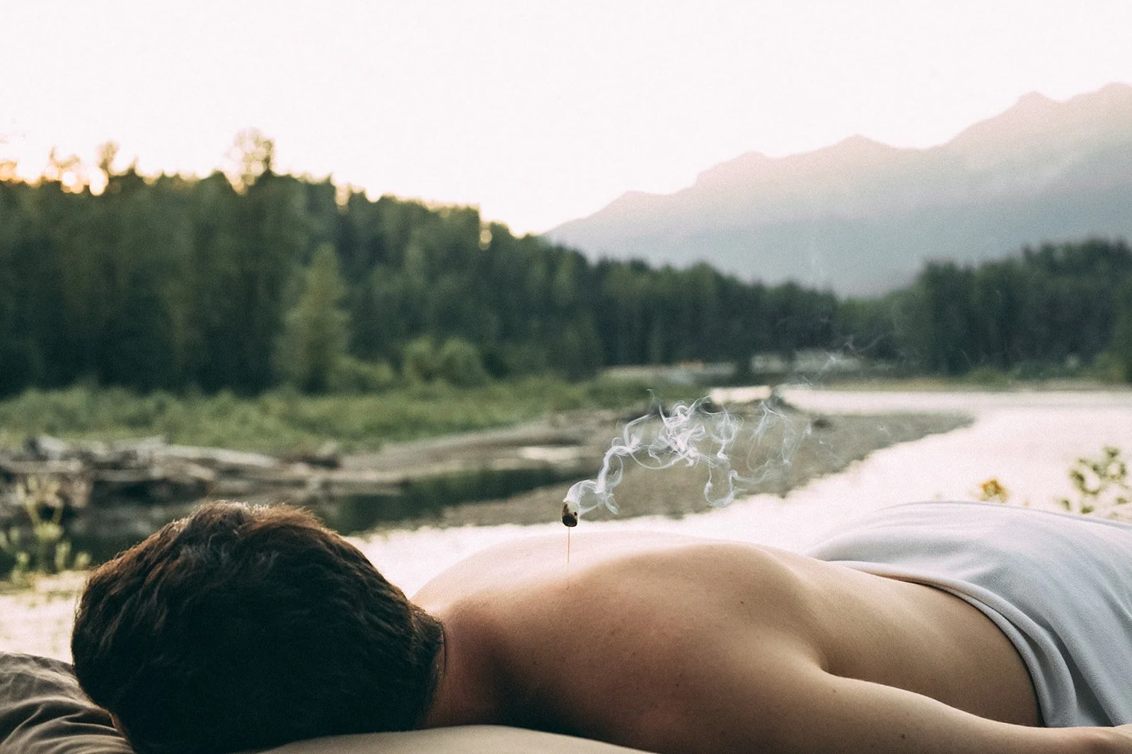 Person lying face down outdoors near a river, with mountains and trees in the background, and a lit incense stick with smoke rising from it on their back.