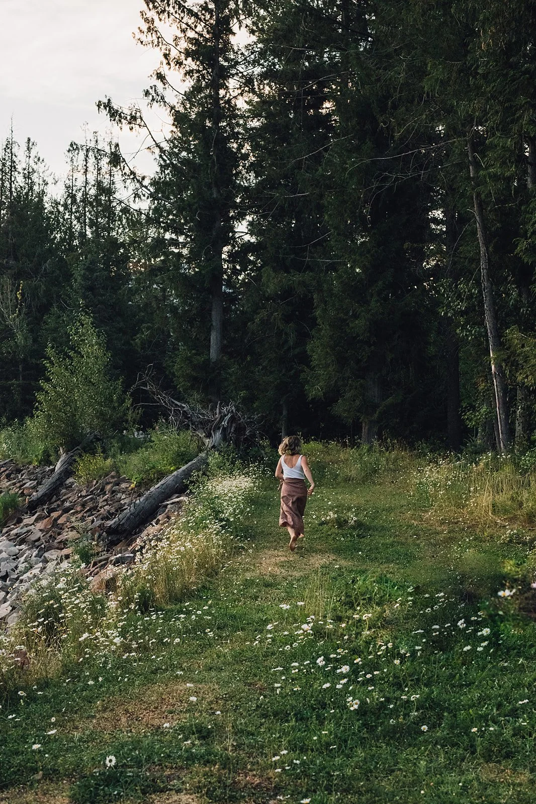 A woman walking along a narrow dirt trail surrounded by wildflowers, with a forest of tall trees in the background.