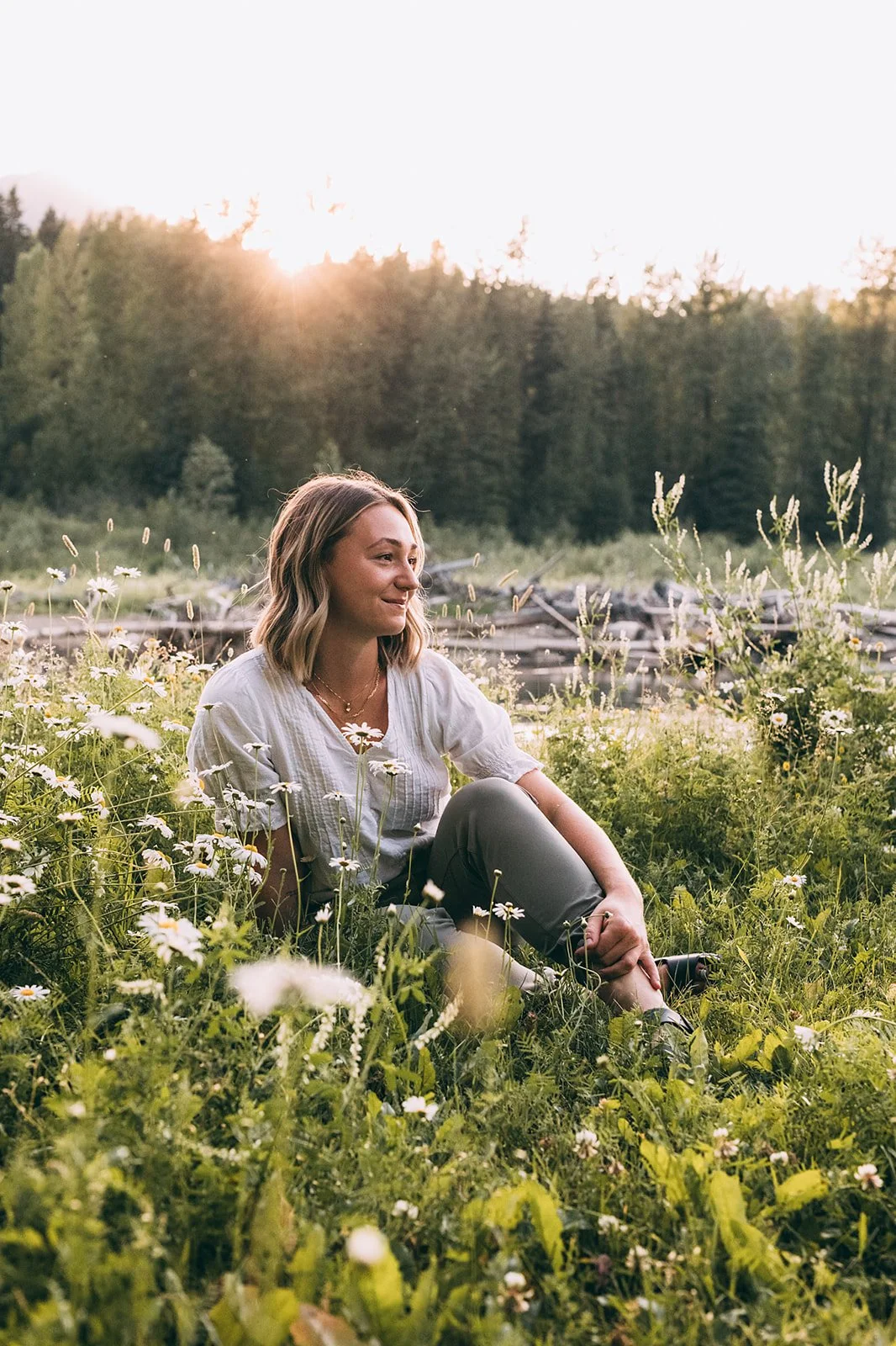A woman sitting in a field of wildflowers near a lake, with trees and a sunset in the background.