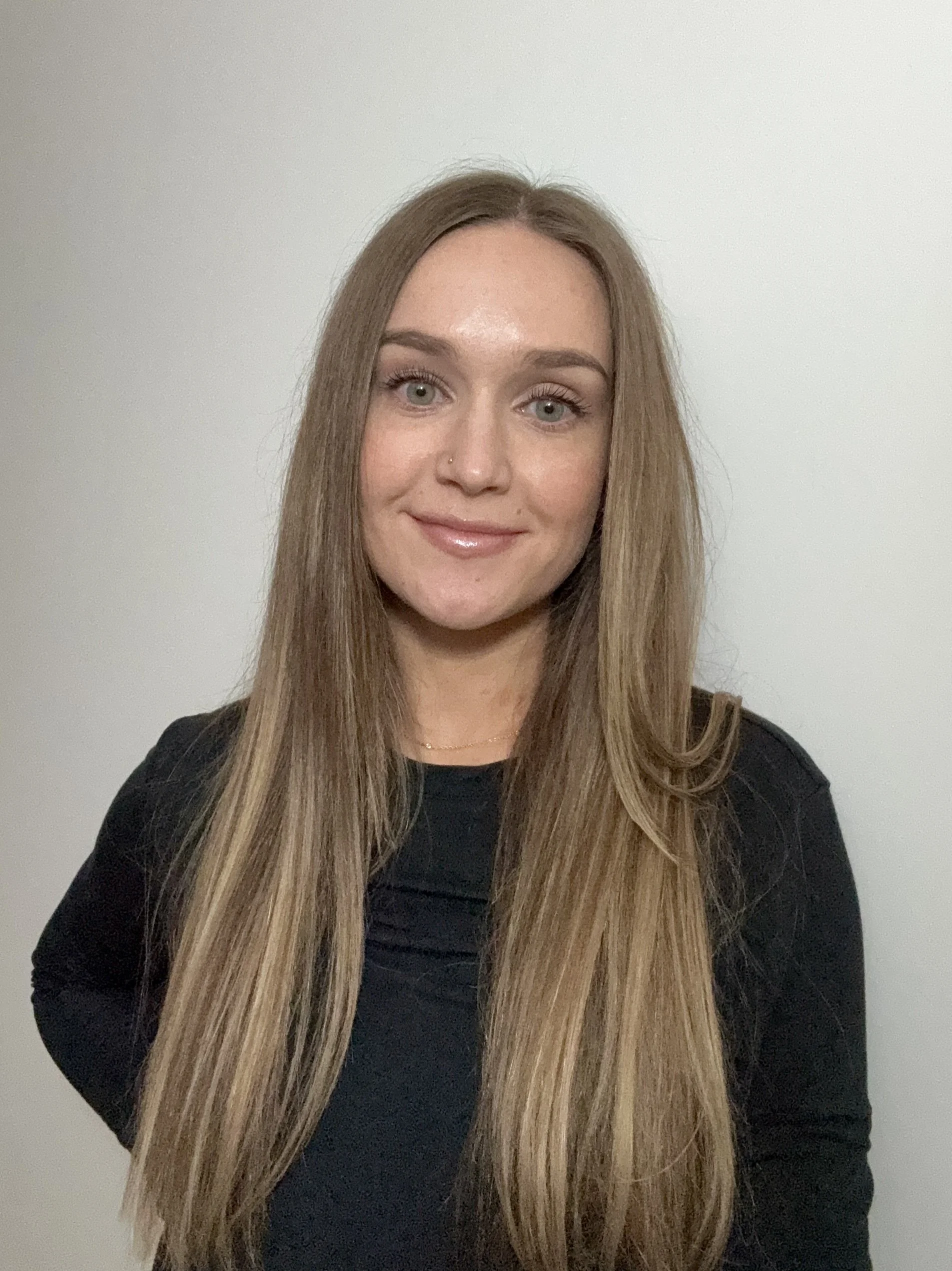 A woman with long straight blond hair wearing a black top, standing against a plain white wall, smiling softly at the camera.
