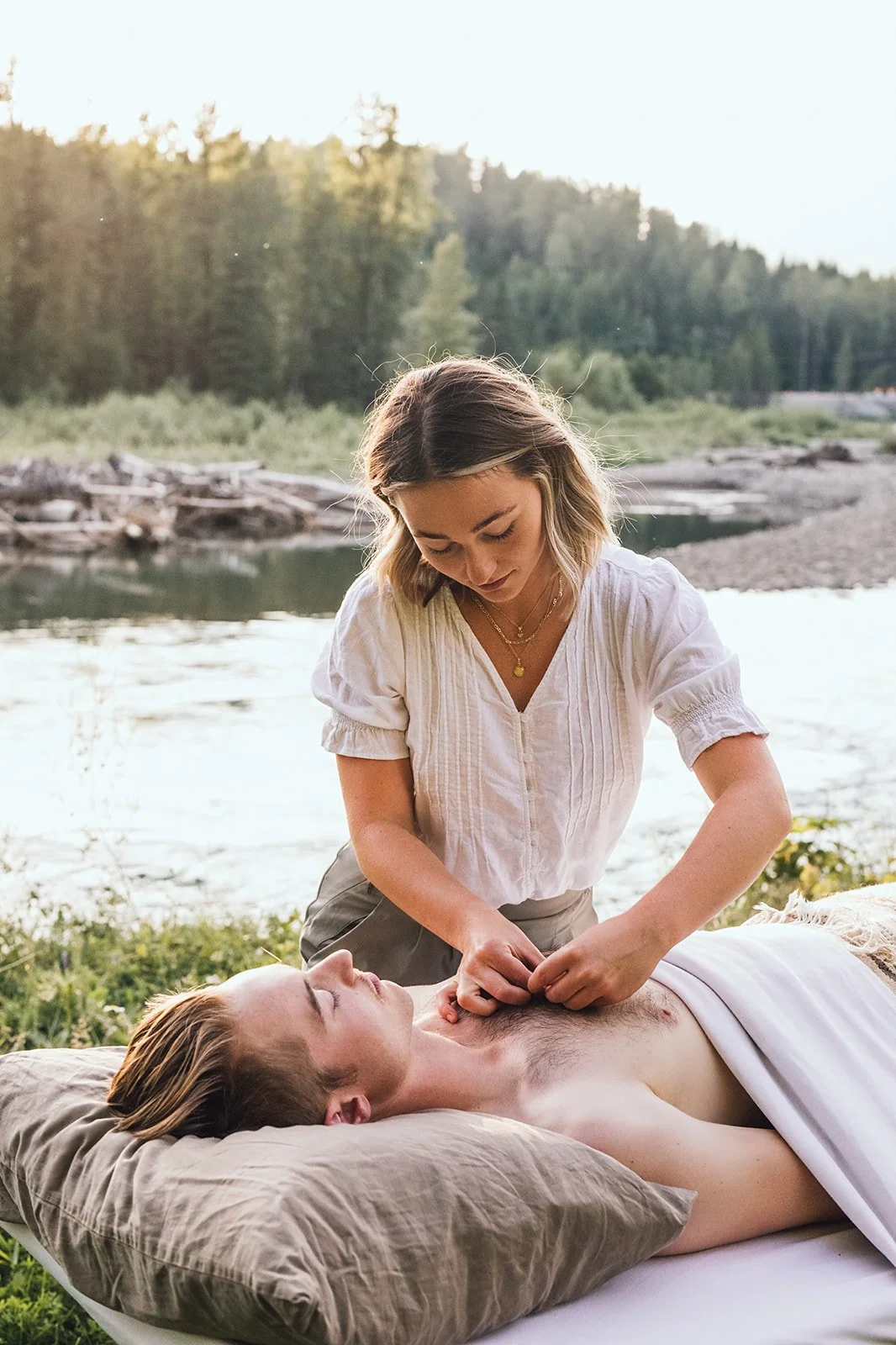 A woman giving a massage to a man lying outdoors near a river, with a forest in the background during sunset.