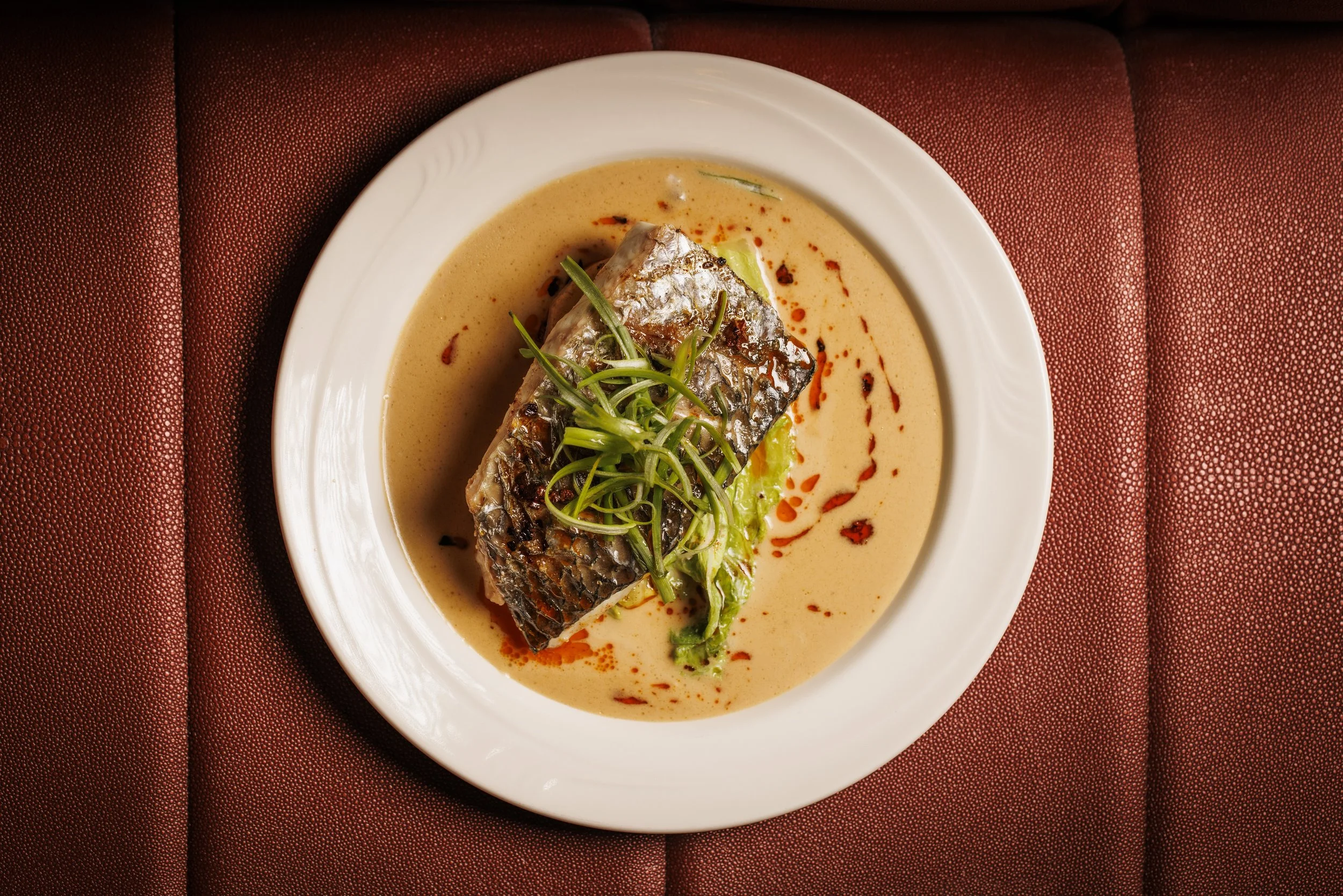 A plated dish featuring a piece of fish topped with green onions, served in a creamy sauce with a drizzle of red chili oil on a white plate.