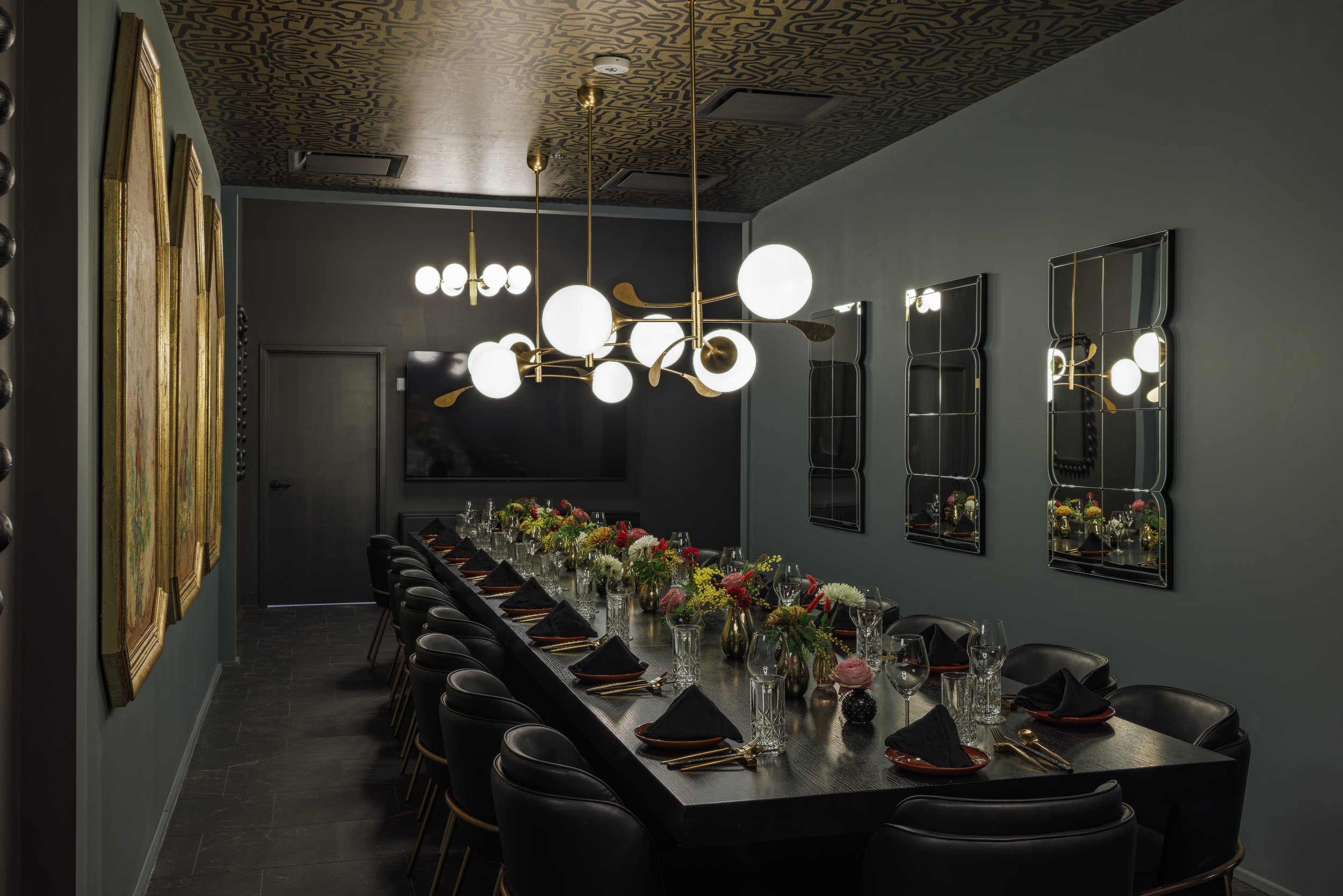 A formal dining room with a long table set for a meal, decorated with flowers and glassware. There are black chairs, modern mirrors on the wall, and contemporary lighting fixtures.
