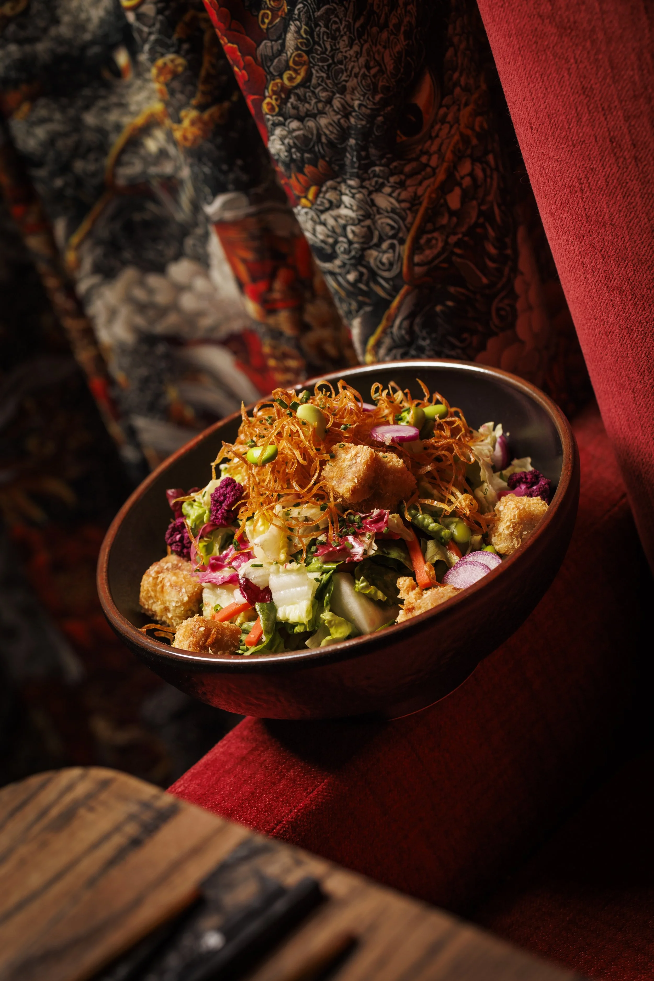 A bowl of mixed salad with lettuce, shredded carrots, radish slices, croutons, and shredded cheese, placed on a red fabric surface next to colorful patterned fabric.