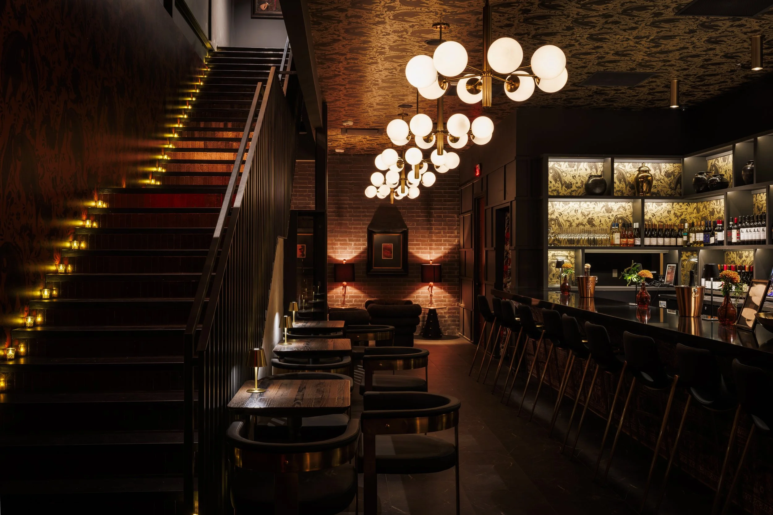 Interior of a dimly lit bar or lounge with a staircase on the left, modern chandeliers hanging from the ceiling, a brick wall with lamps and artwork in the background, a bar counter with barstools on the right, and shelves filled with bottles and gla