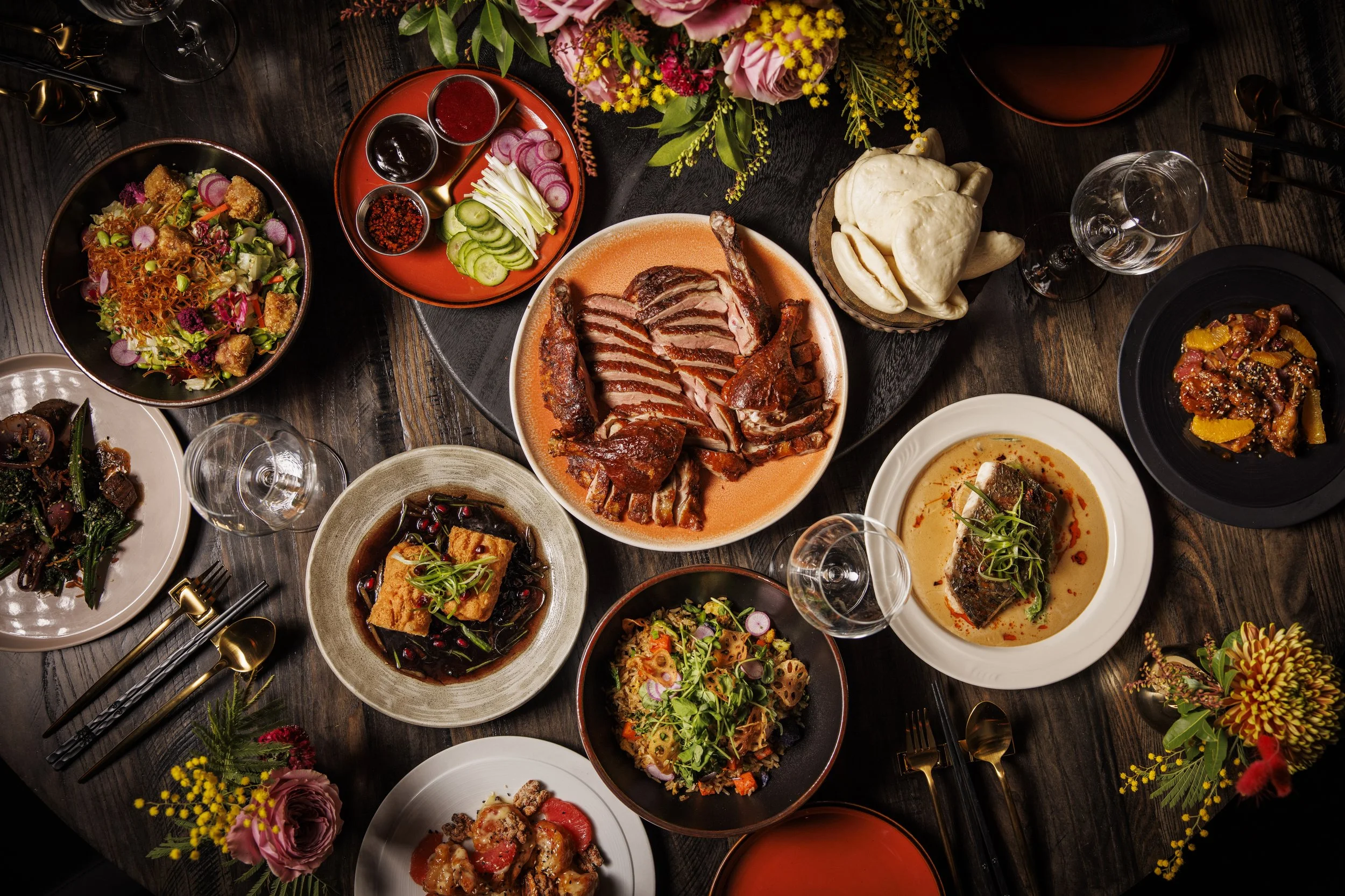 A variety of Asian dishes served on a dark wooden table with floral decorations. The dishes include sliced roasted duck, salad, tofu, fish in sauce, spicy beef, vegetables, and condiments.