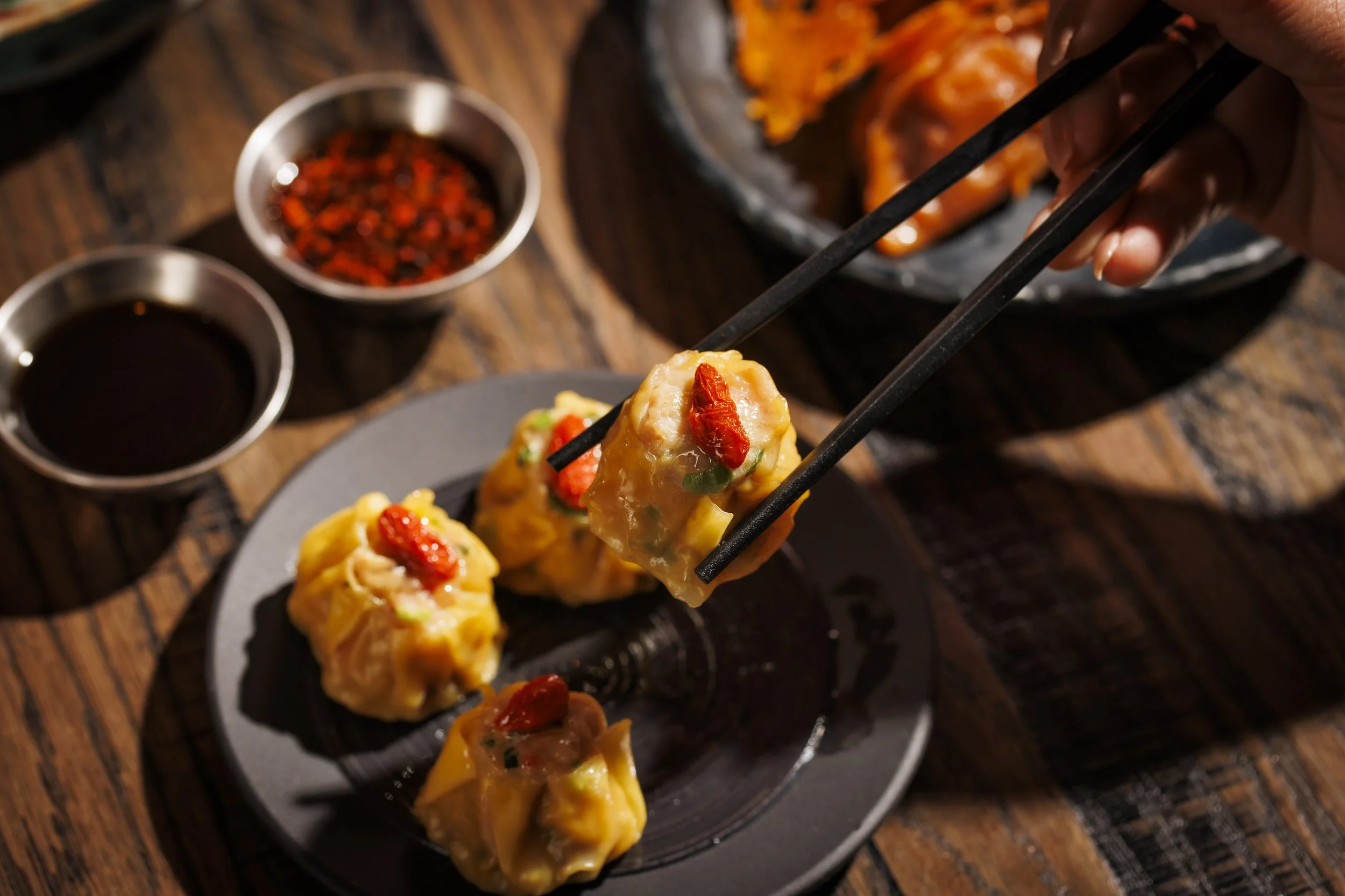 Asian-style dumplings with red chili topping, served on a black plate with dipping sauces and a hand using chopsticks to pick up a dumpling, on a wooden table.