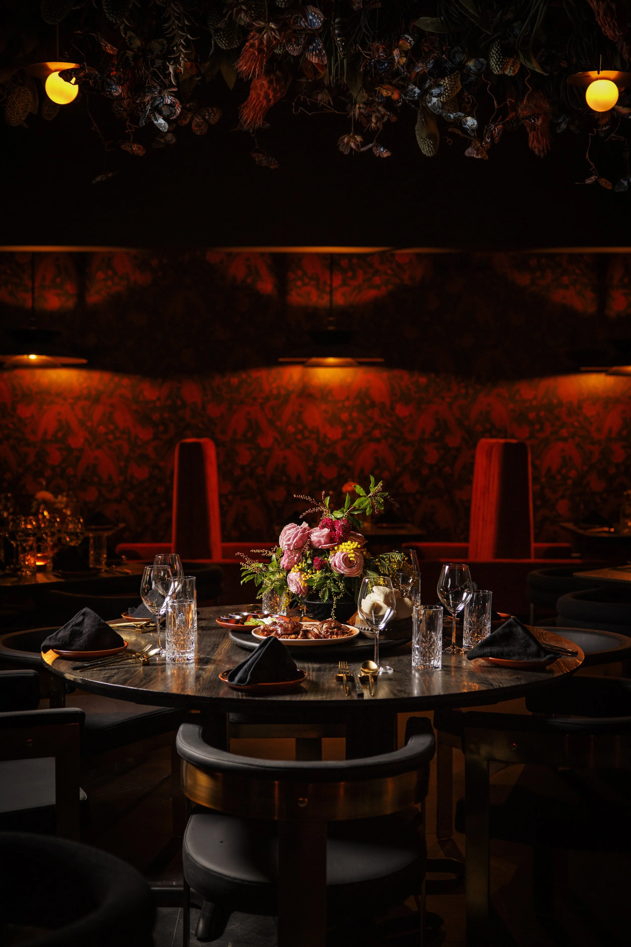 Elegant dining table set with black napkins, glassware, and a floral centerpiece in a dimly lit restaurant with red and black decor.