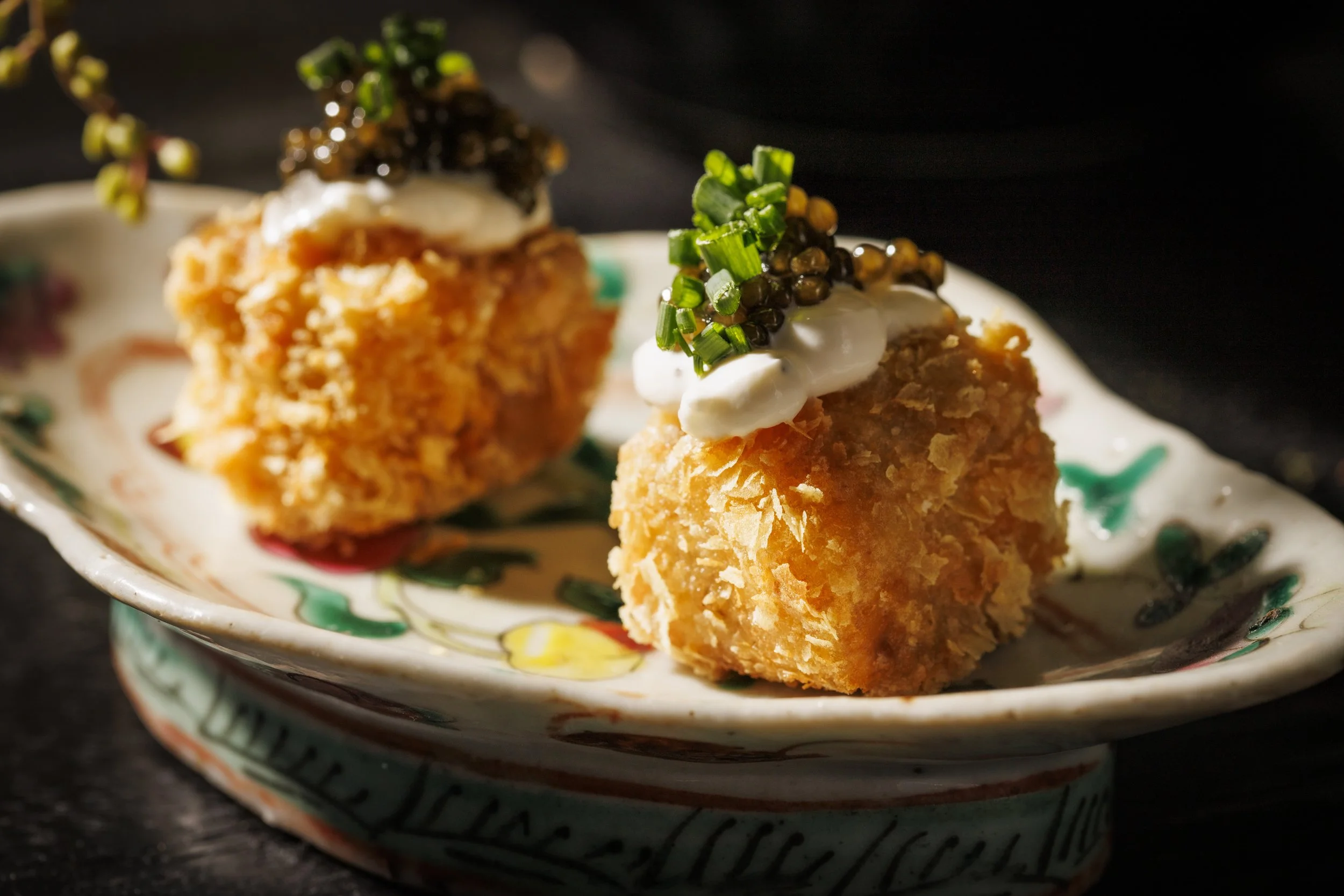 Two deep-fried golden-brown croquettes topped with sour cream, chopped chives, and black caviar, served on a decorative ceramic plate.
