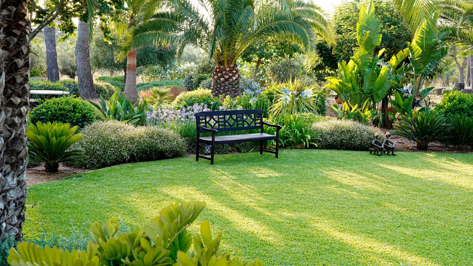 A lush garden with a neatly maintained green lawn, surrounded by various vibrant plants and trees. There's a black wooden bench in the center, and decorative sculptures and blooming flowers are visible in the background.